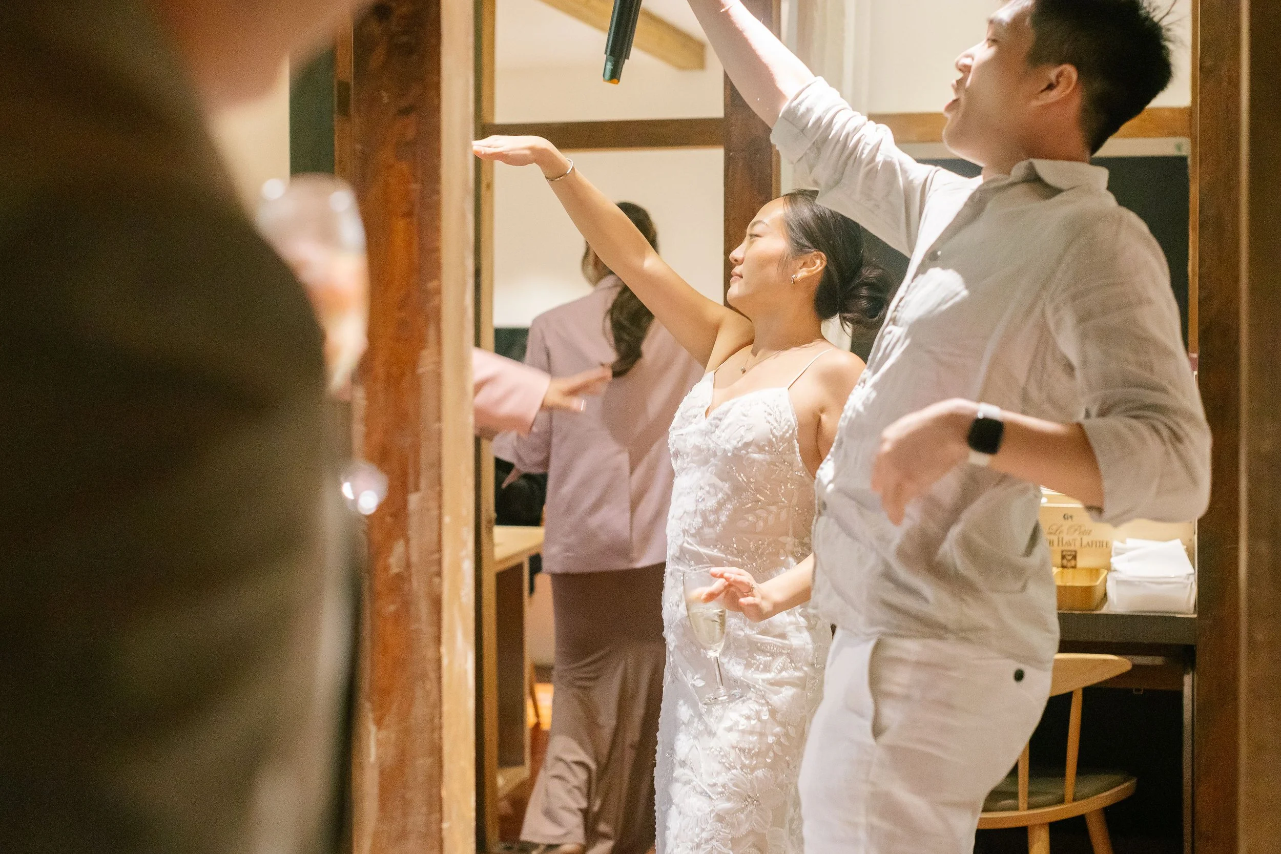 A woman in a white wedding dress is raising her arm with a glass in her hand, accompanied by a man in a white shirt, at an event with others in the background.