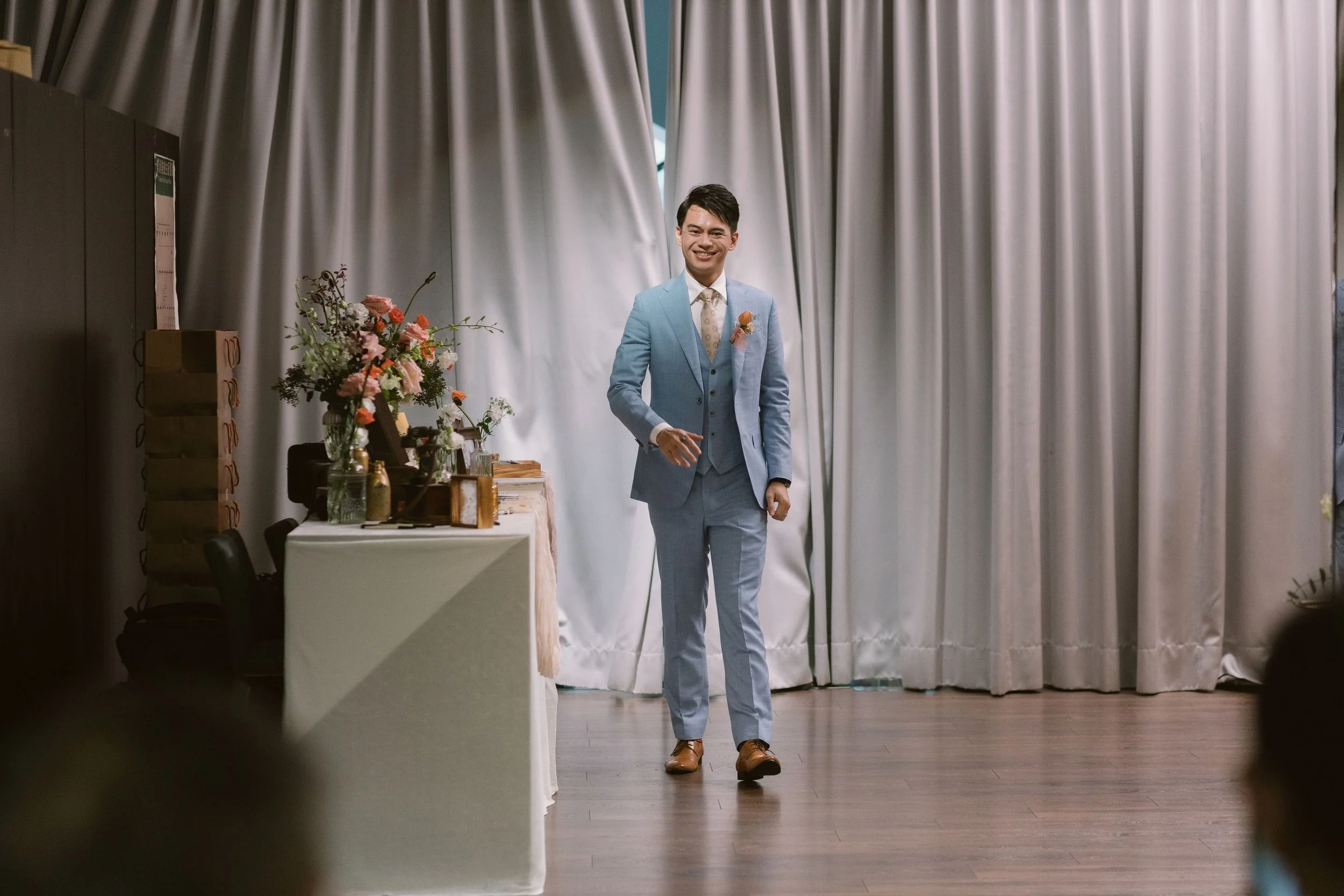 A man dressed in a light blue suit and brown shoes walking in front of a beige curtain, with a table decorated with flowers and a vase on his left.
