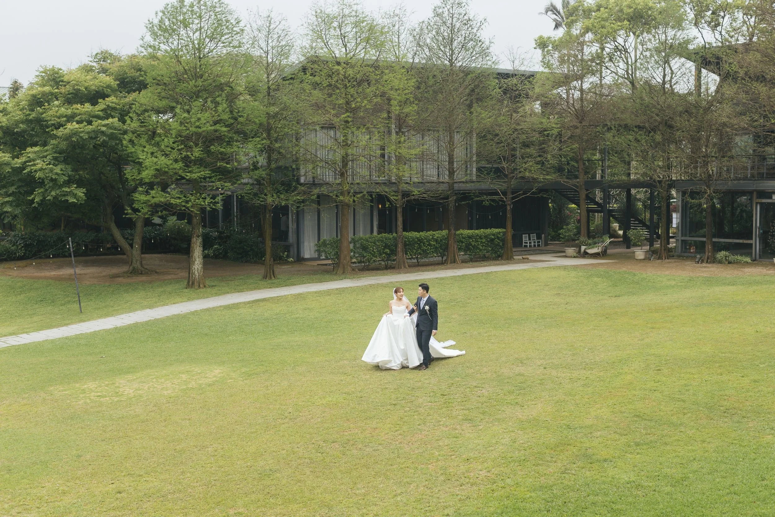 A bride and groom in wedding attire walking outdoors on a grassy area surrounded by trees and modern buildings in the background.