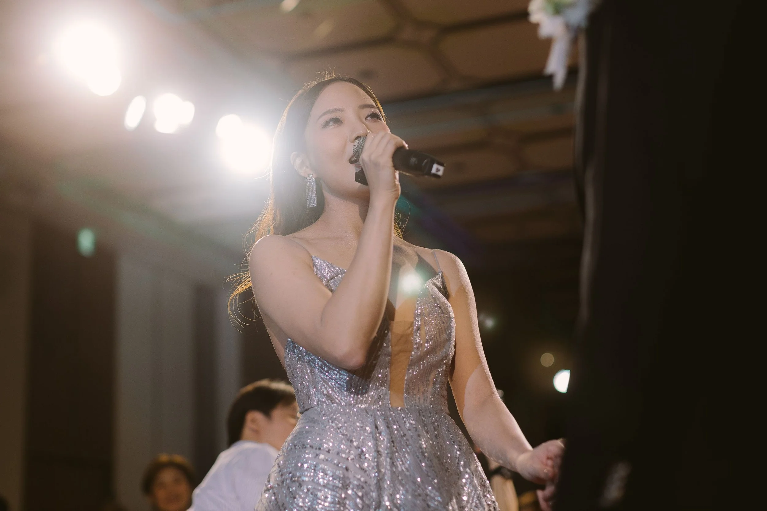 A woman in a sparkly dress singing into a microphone at an indoor event with bright lights.