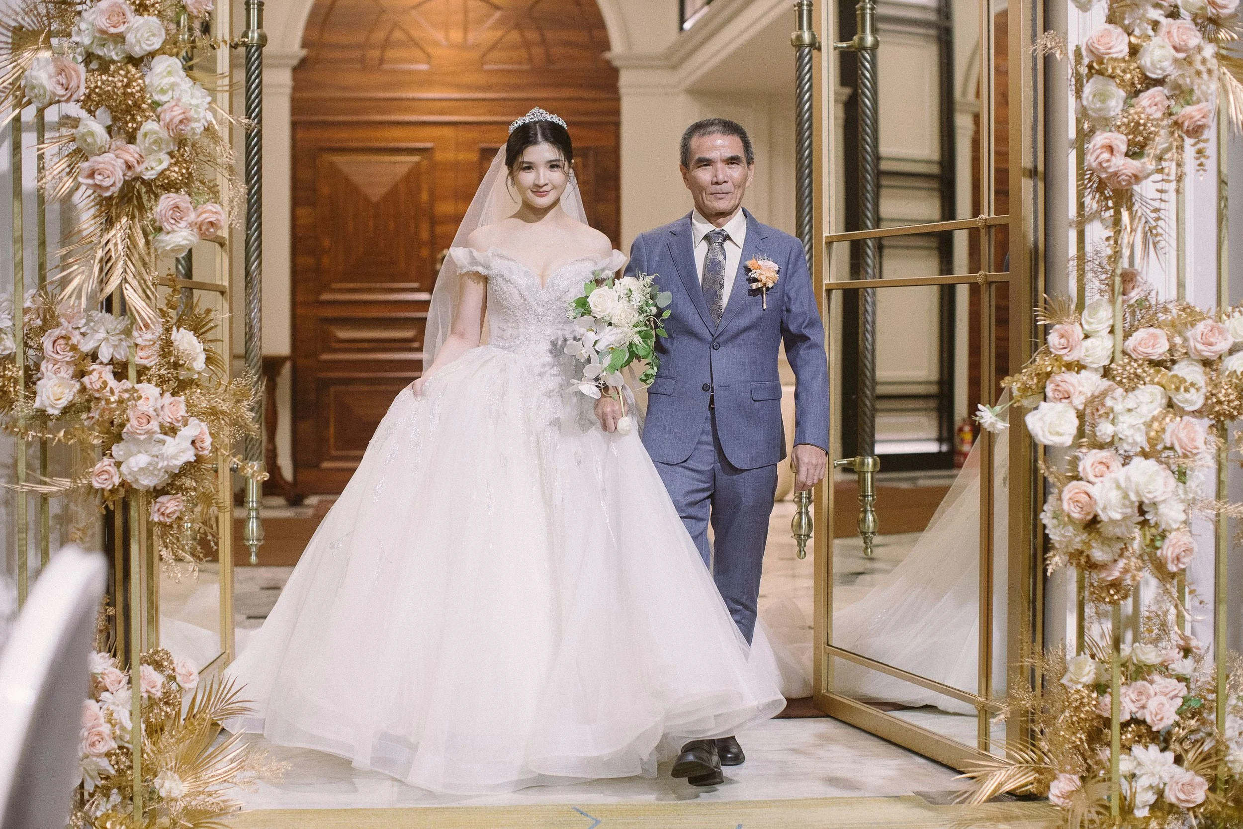 A young woman in a white wedding gown and veil standing beside an older man in a blue suit, both smiling, in a decorated wedding venue with floral arrangements on either side of them.