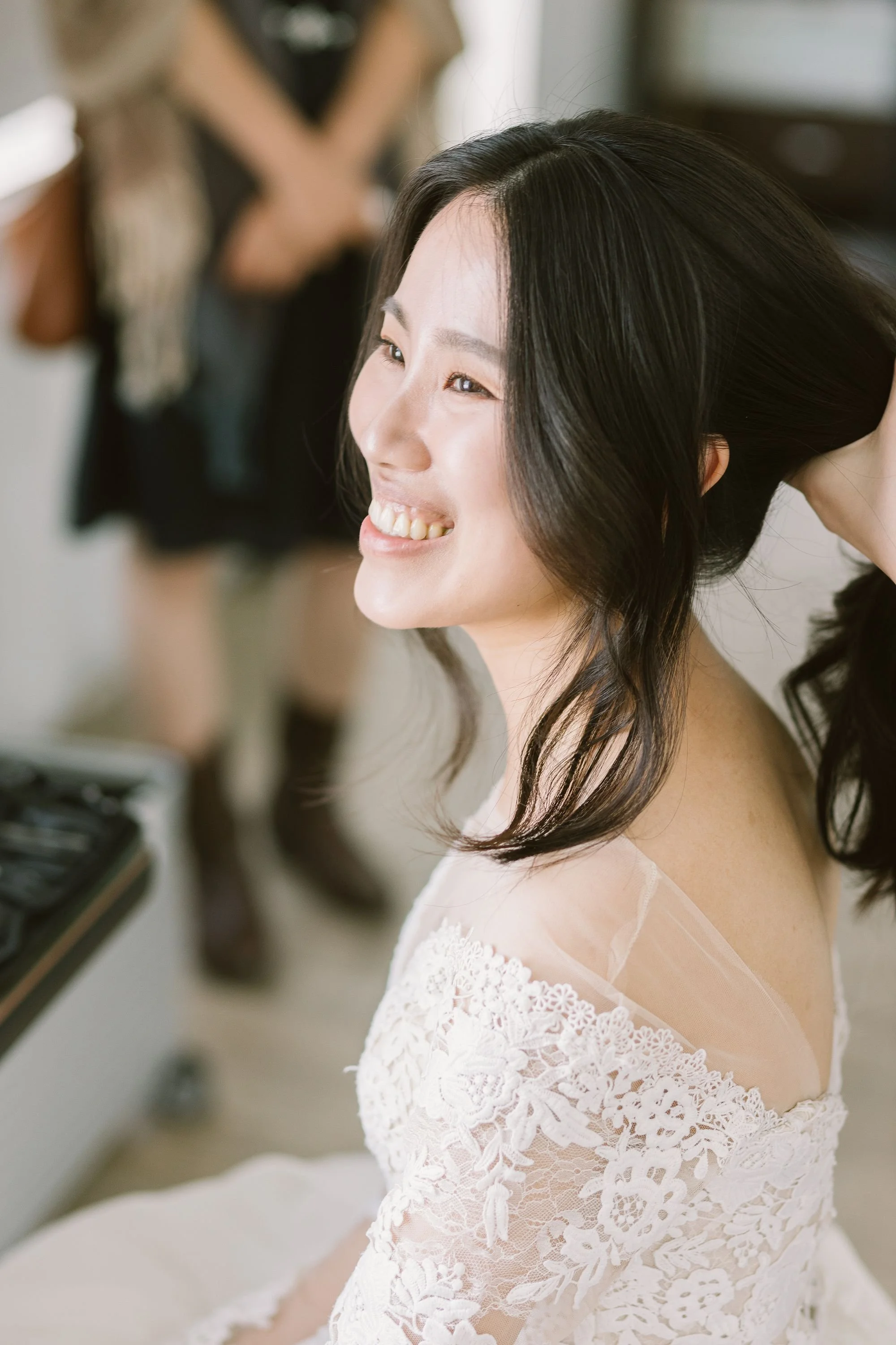 A woman with long dark hair smiling as she gets ready in a white lace wedding dress.