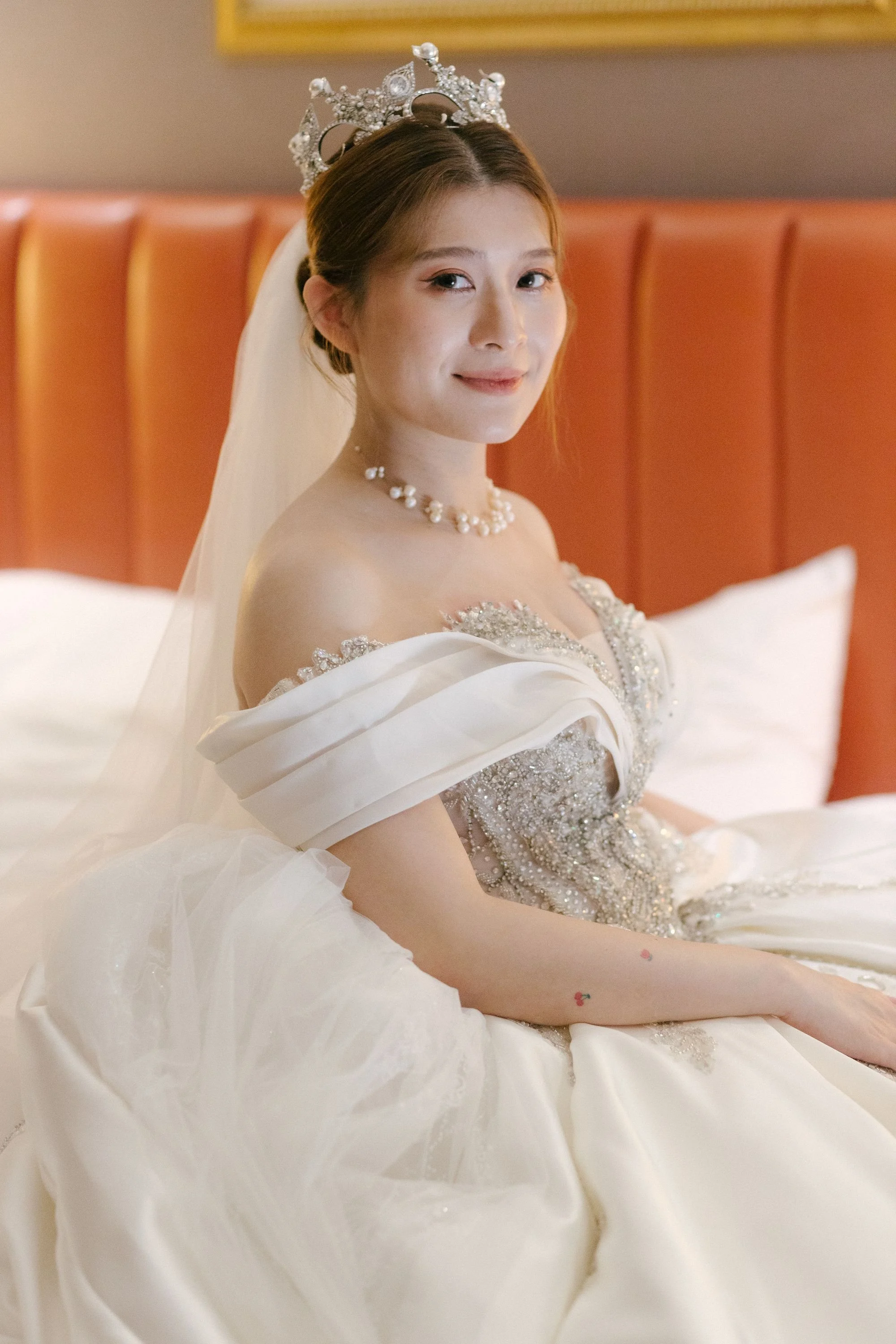 A young woman in a wedding dress wearing a silver tiara and pearl necklace, sitting on a bed with an orange headboard and smiling gently.