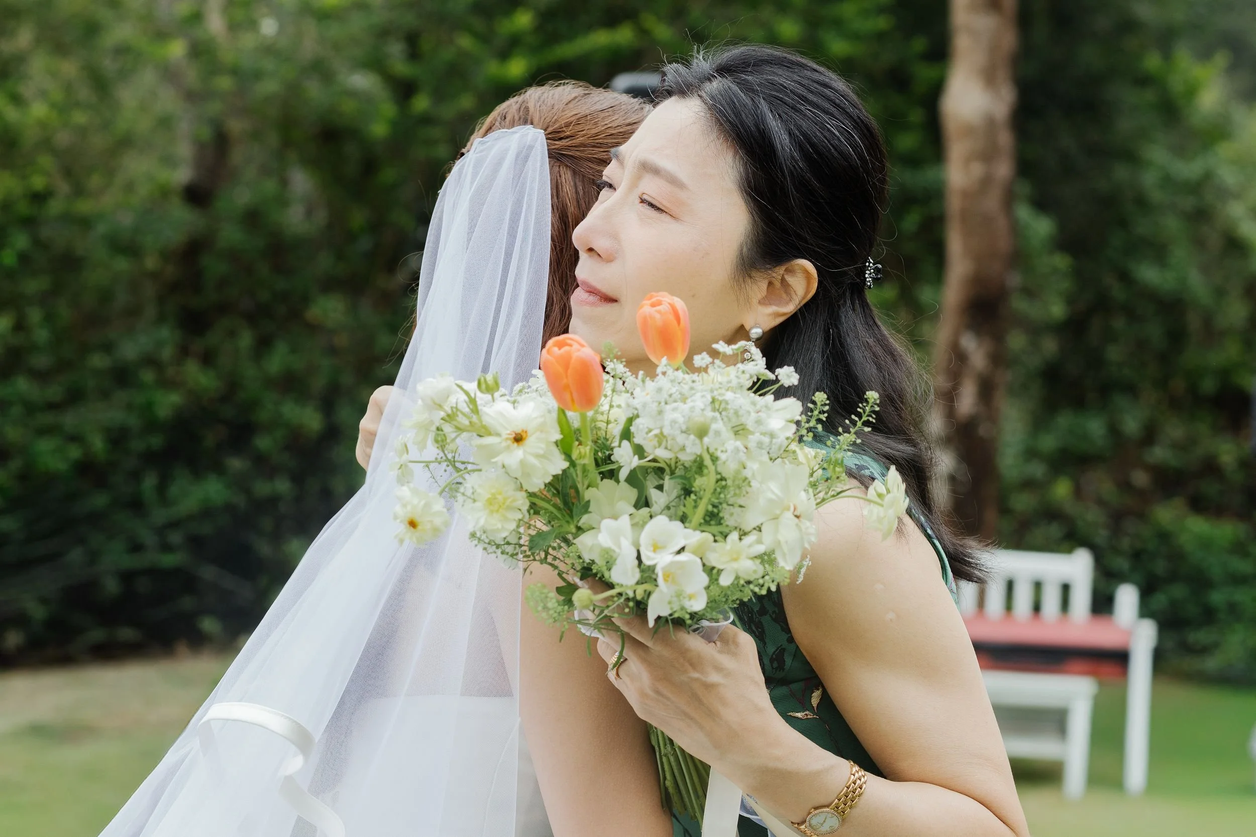 A woman with black hair, earrings, and a gold watch holding a bouquet of white and orange flowers, embracing another woman with a veil in an outdoor garden setting.