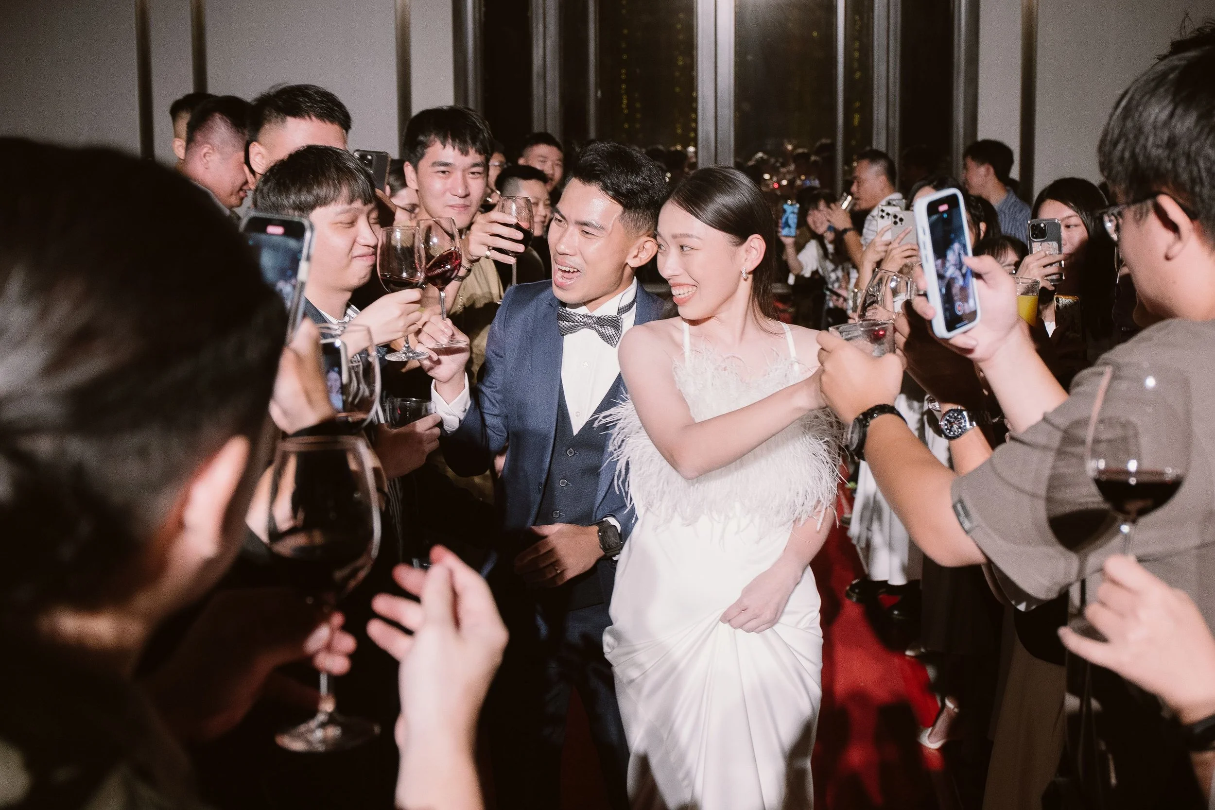 A bride and groom celebrating with friends at a wedding reception, surrounded by guests holding up glasses and taking photos.