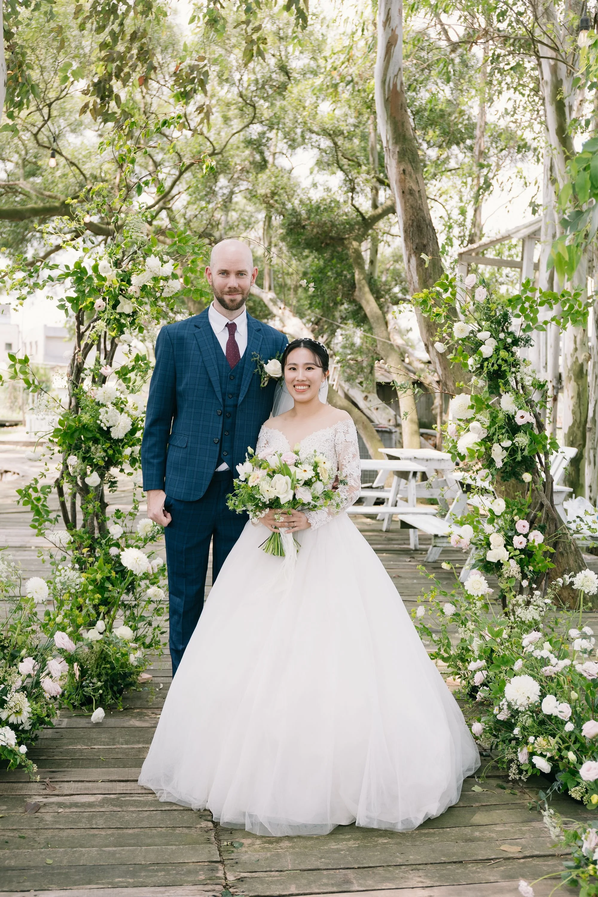 A newlywed couple standing under a floral archway outdoors on a wooden deck. The groom is tall, bald, with a beard, wearing a blue plaid suit with a white shirt and red tie. The bride is shorter, with dark hair, wearing a white wedding gown with lace