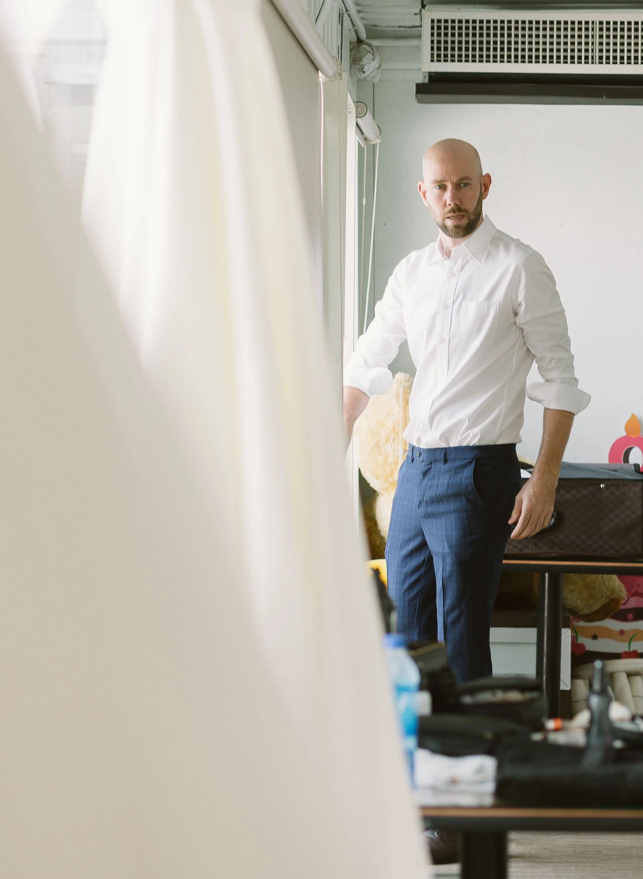 A man with a shaved head and beard wearing a white shirt and dark blue plaid pants, standing in a room with various objects and a white wall in the background.