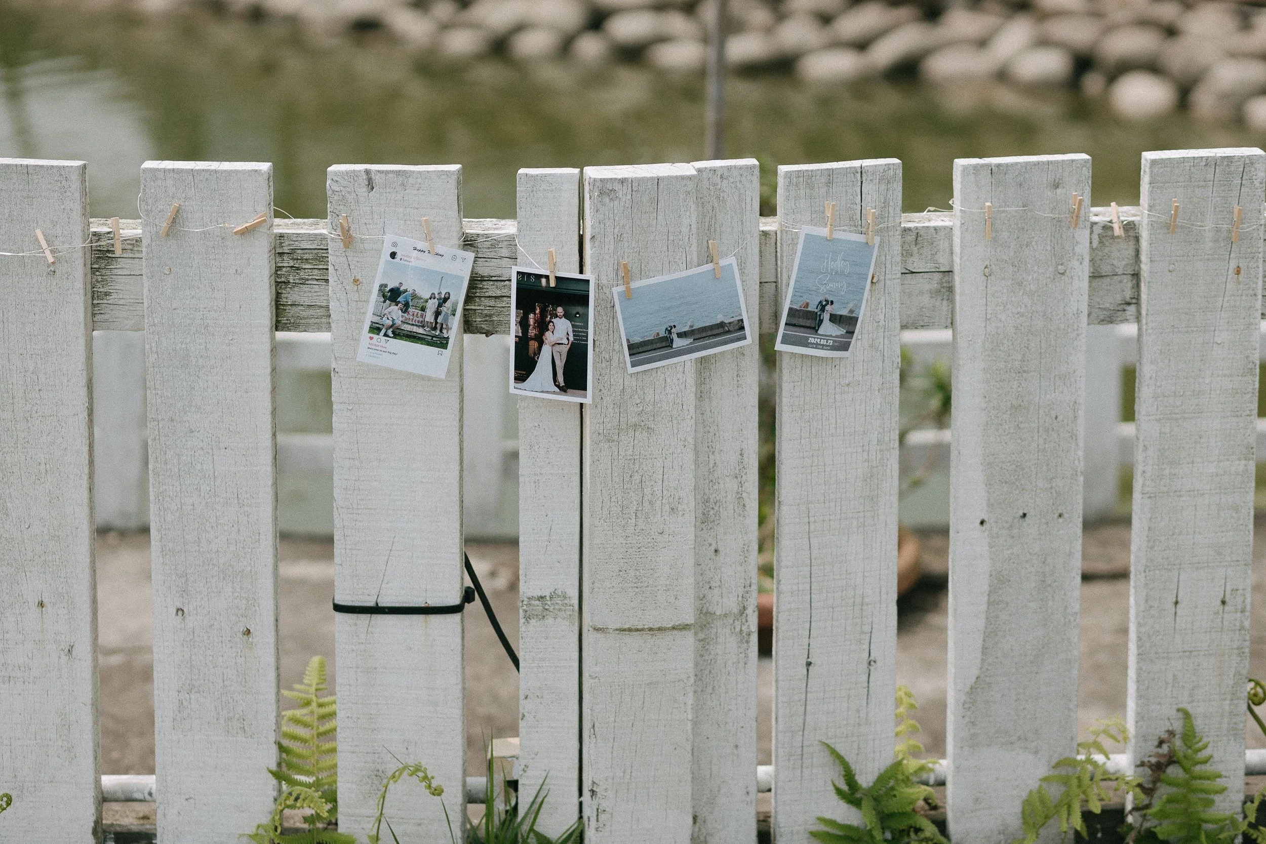Photographs hanging on a white wooden fence with clothespins, overlooking a body of water with rocks in the background.
