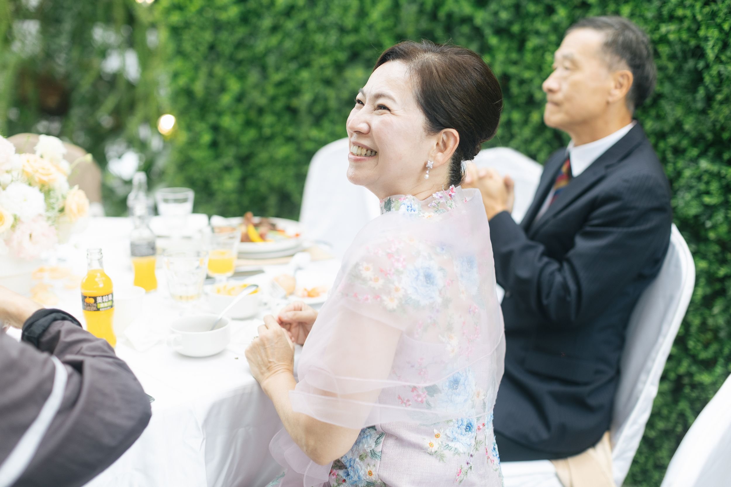 Elderly woman smiling and seated at a table with others during an outdoor meal or celebration, with greenery in the background.