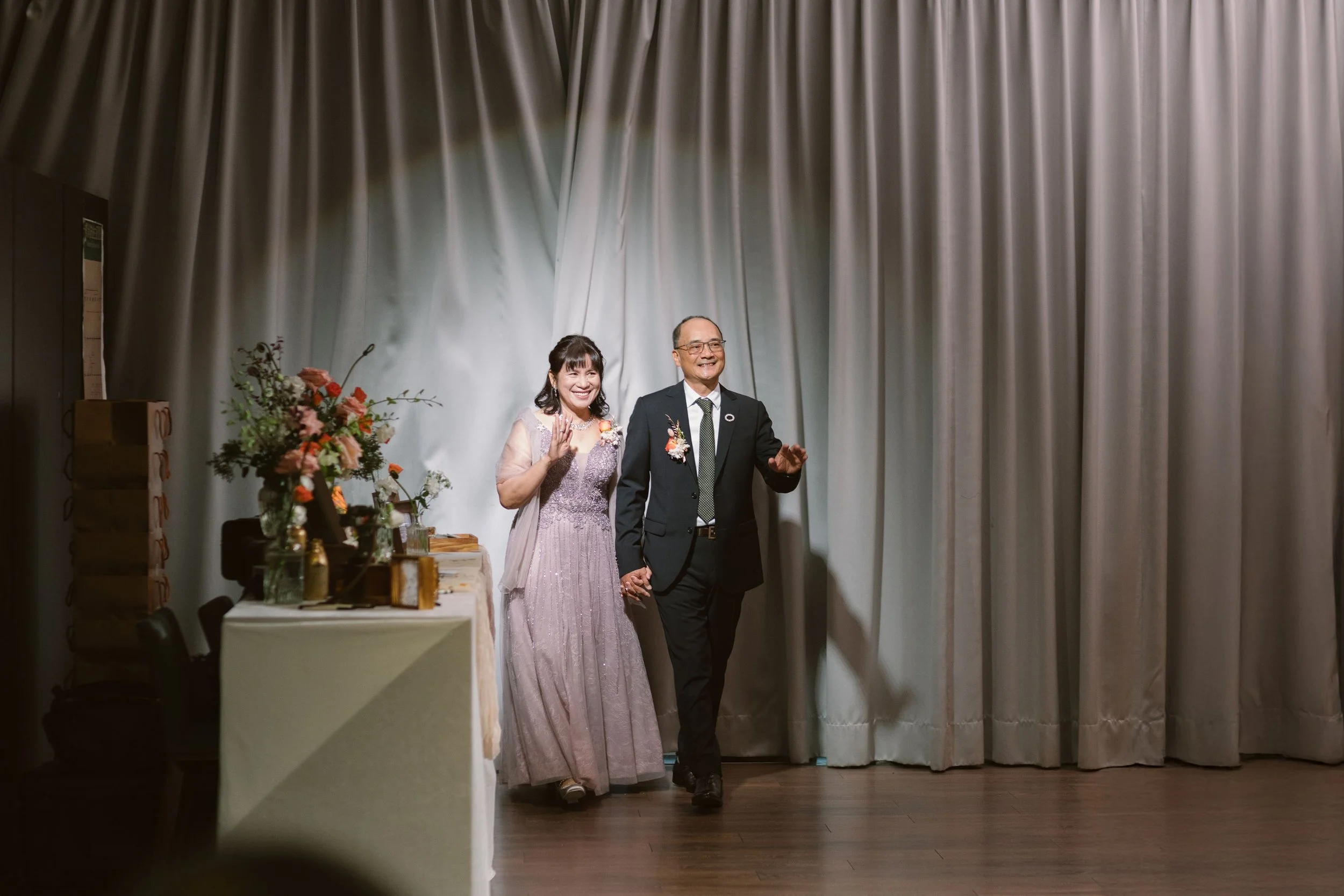 A couple dressed in formal attire holding hands and smiling on stage, with a curtain background and floral decorations to the side.