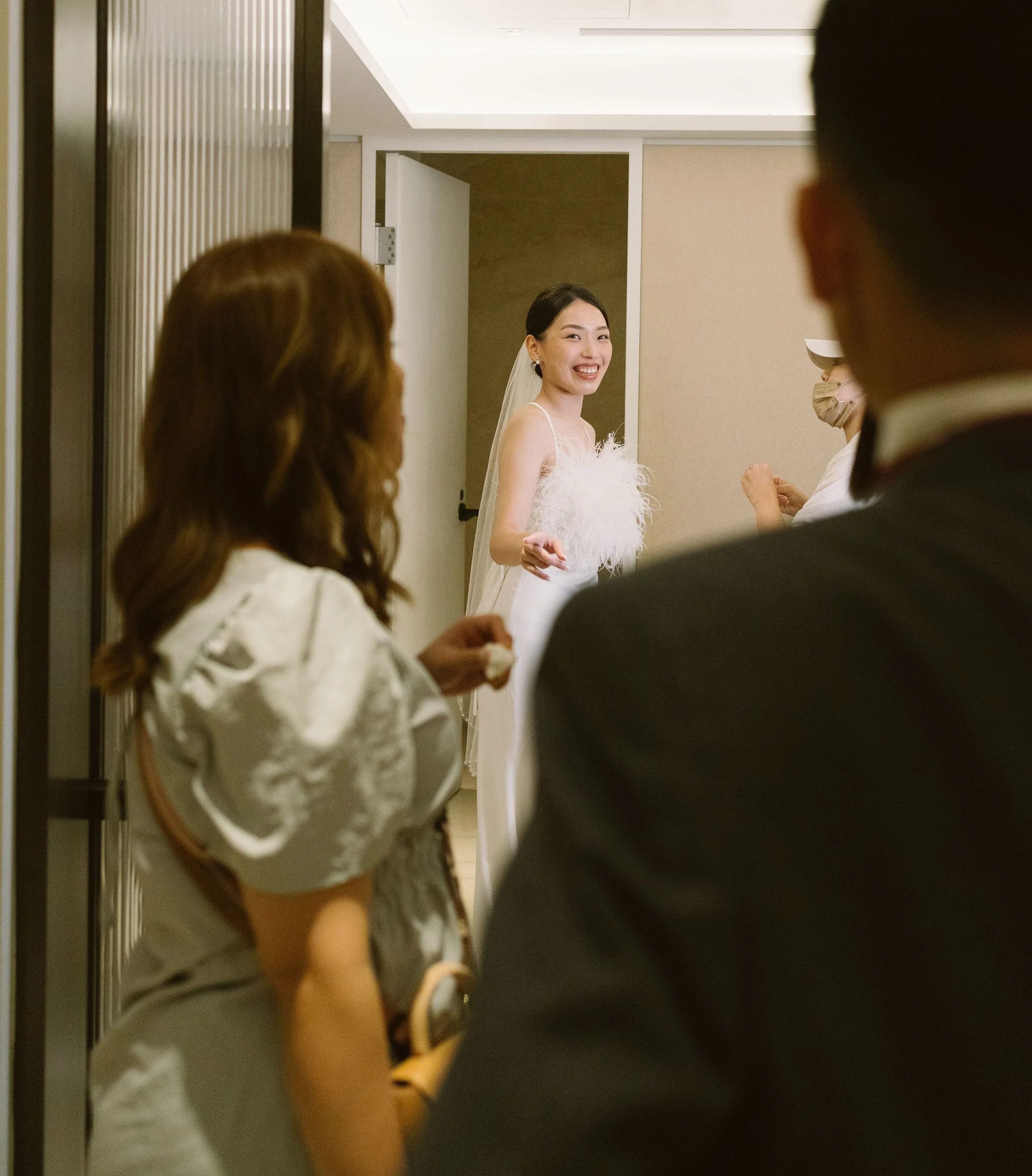 A happy bride smiling and looking at the camera inside a hotel or wedding venue, with guests partially visible in the foreground.