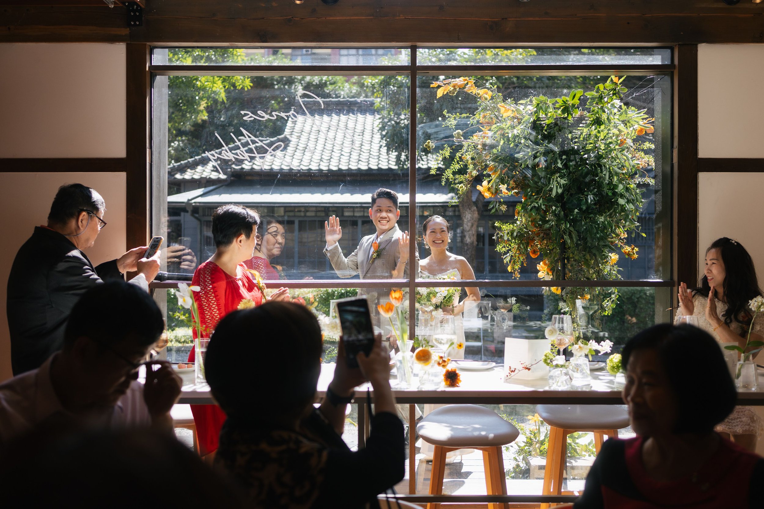 A wedding celebration viewed through a window, featuring a smiling bride and groom sitting at a decorated table with floral arrangements, surrounded by family and friends.