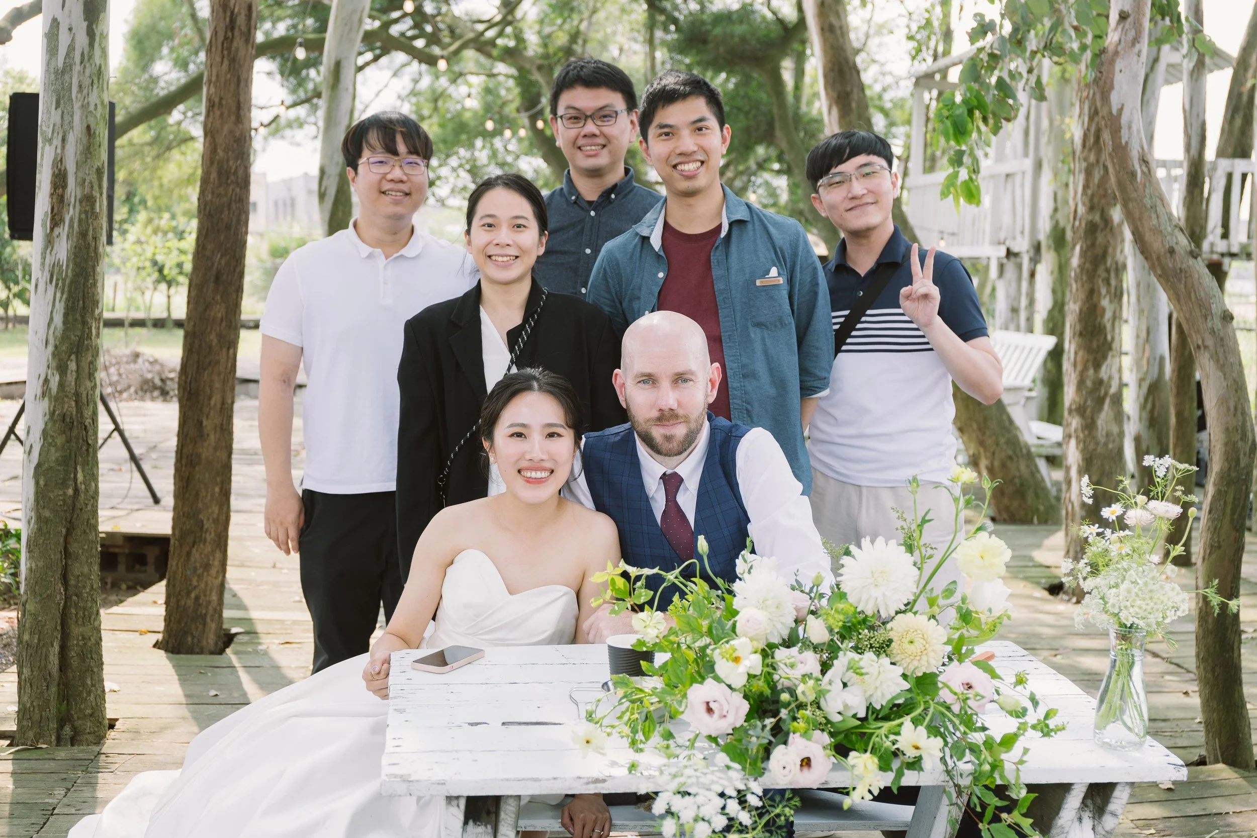 A group of eight people, including a bride in a white wedding dress and a groom in a suit, gathered outside in a wooded area with trees and greenery during a wedding celebration, with a table of flowers in the foreground.