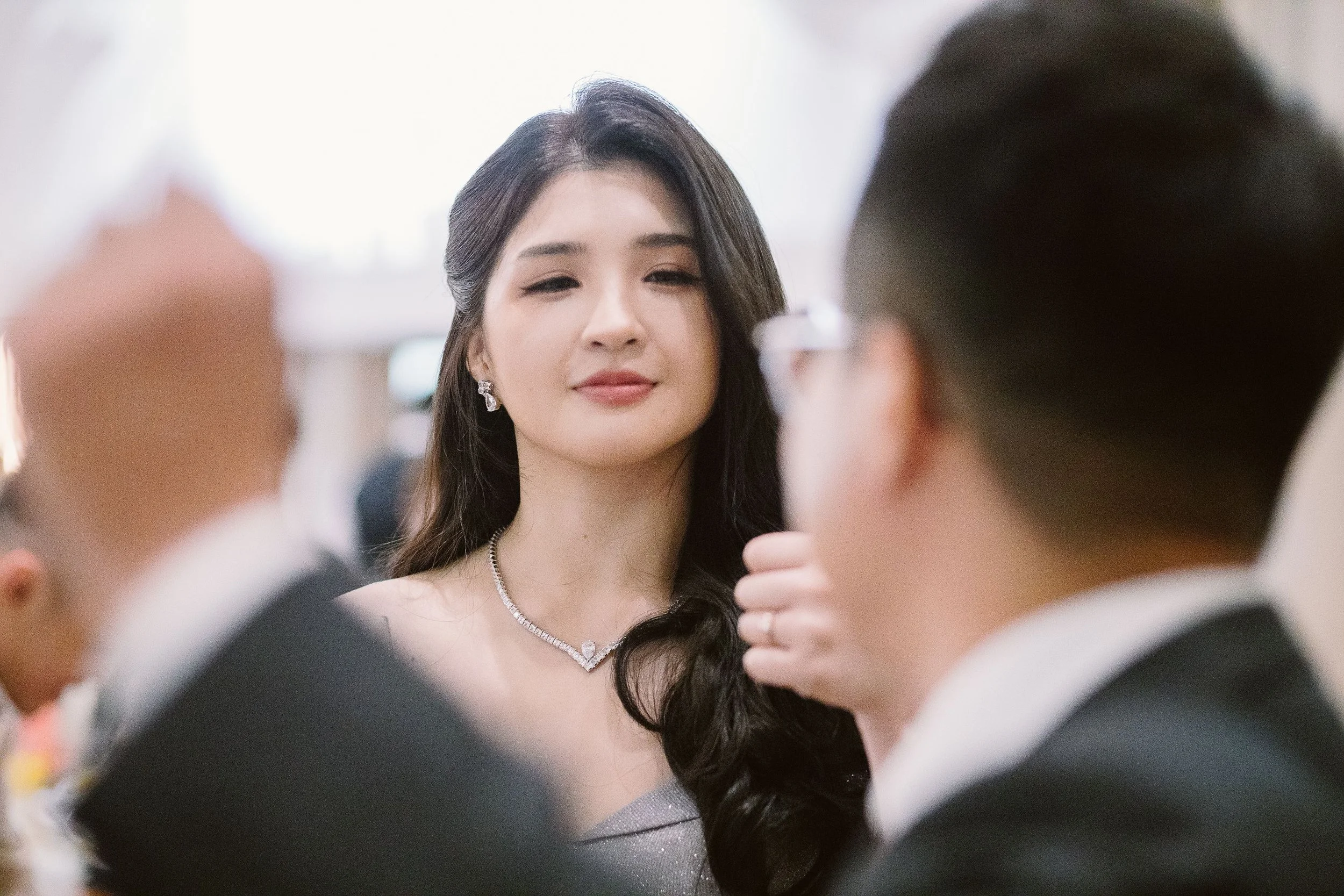 A woman with dark hair and jewelry is being photographed or filmed by two men in suits, with her face centered and the men partially visible in the foreground.