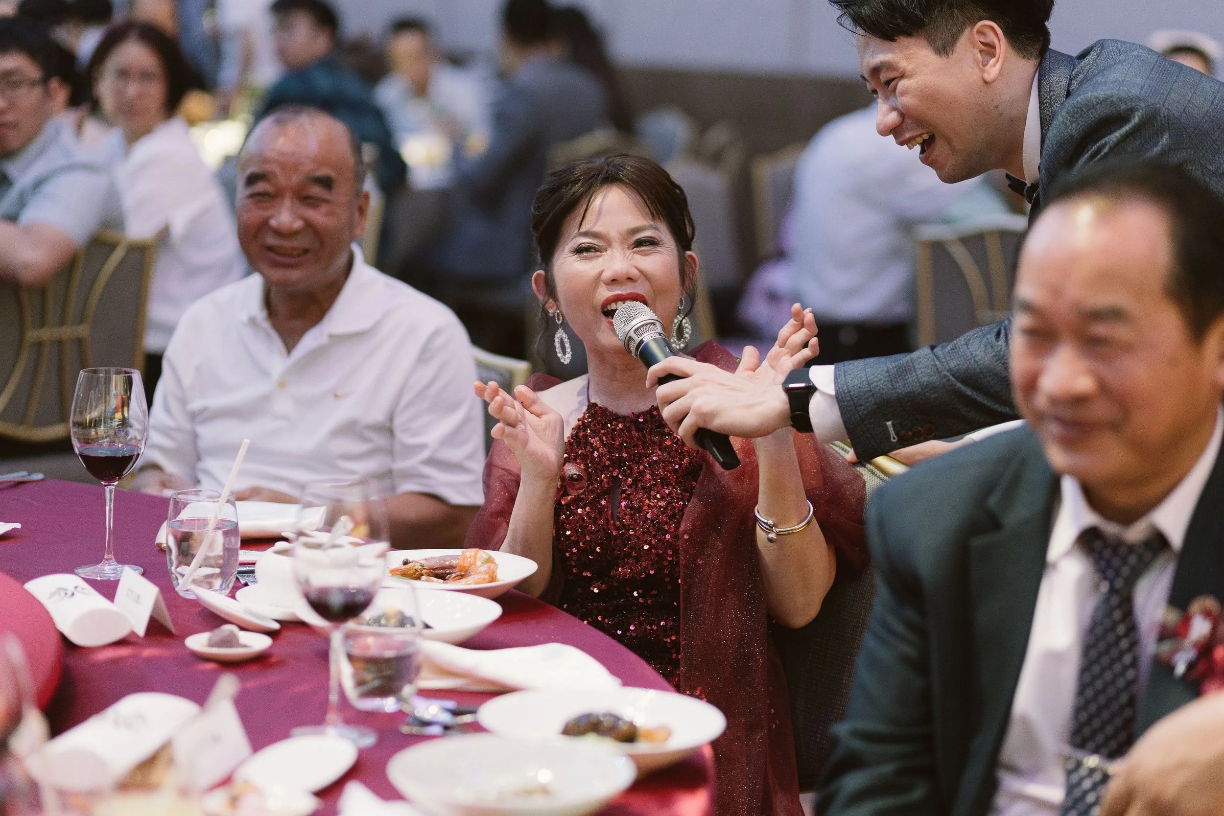 A woman in a red sequined dress is speaking into a microphone, smiling and interacting with a man in a gray suit at a banquet table. The man is leaning in to her, smiling. Other guests are seated at the table, some smiling and talking. There are plat