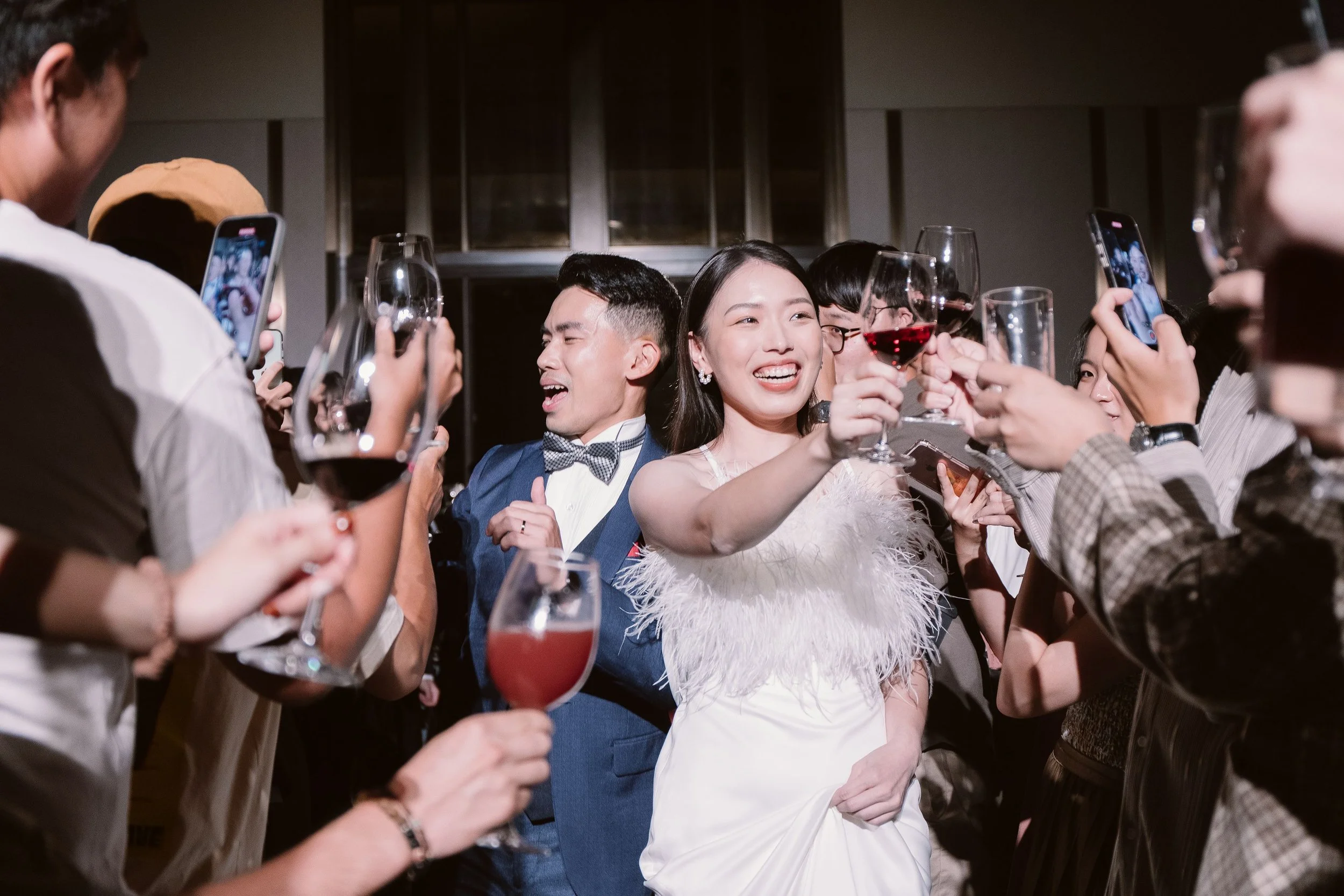 A group of people celebrating at a party, raising glasses of wine and taking photos, with a woman in a white dress smiling at the center.