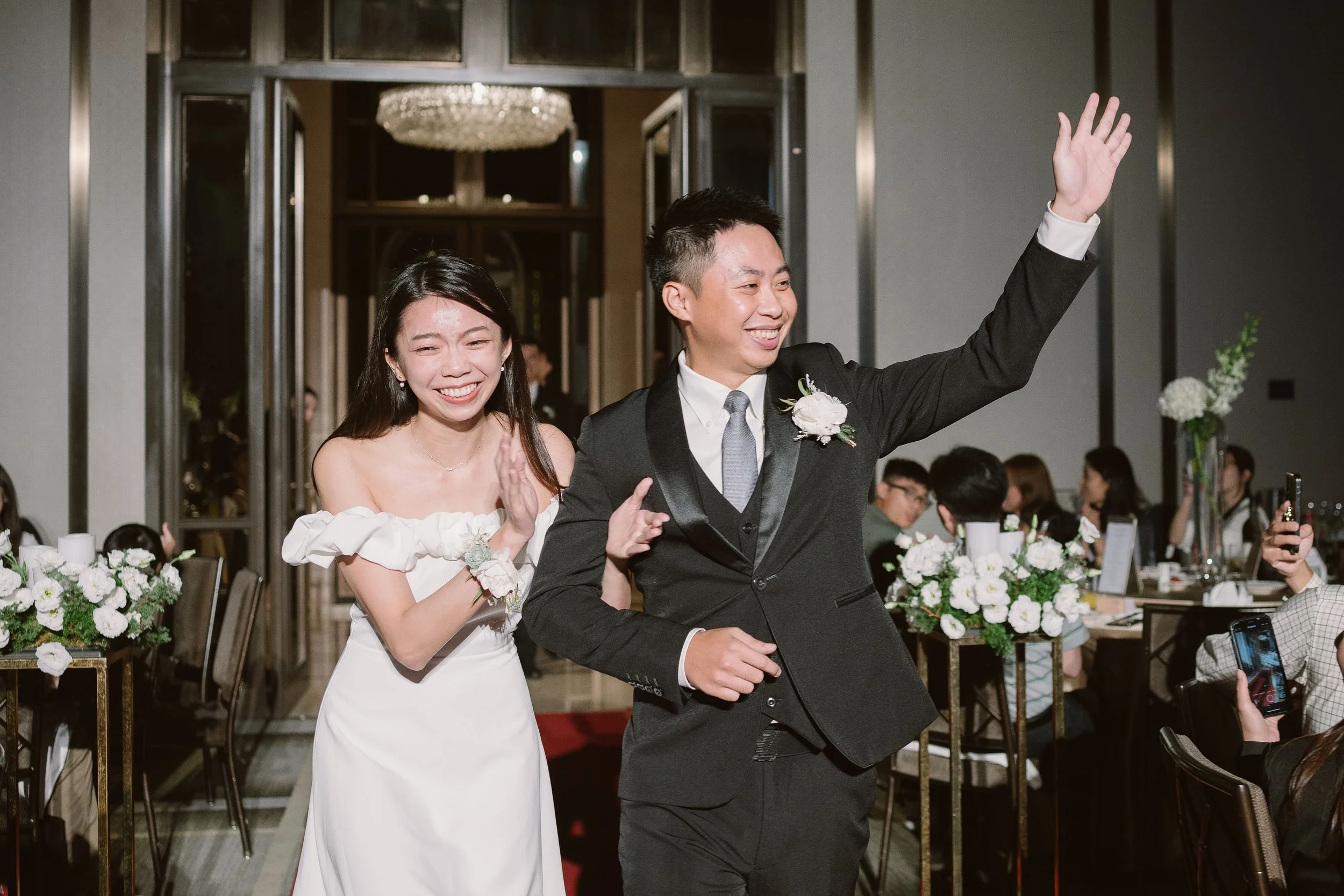 A smiling couple at their wedding reception, with the groom waving. The bride is wearing a white dress with off-the-shoulder ruffles, and the groom is in a black tuxedo with a boutonnière. The reception area is decorated with white flowers and guests