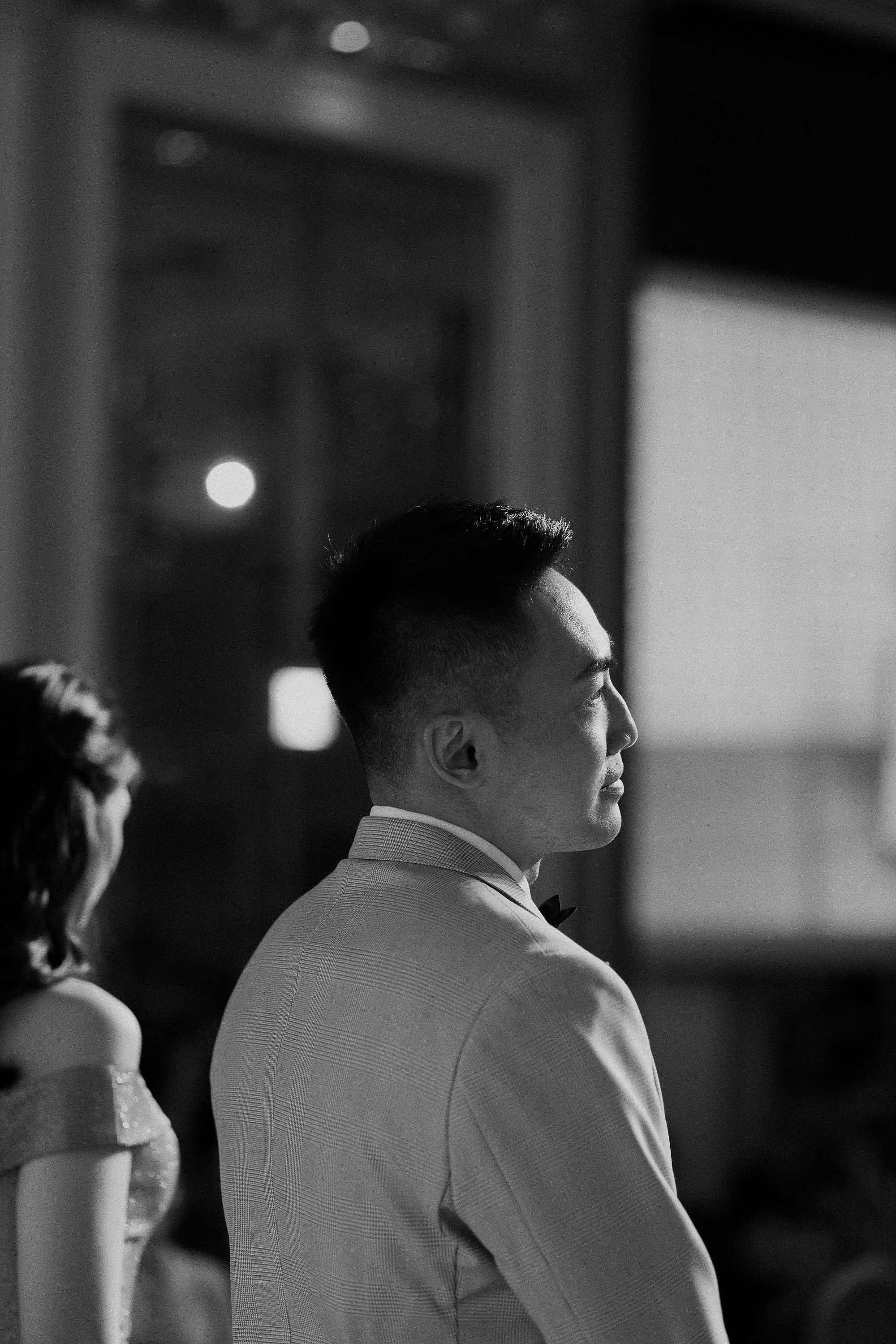 A man in a tuxedo with a bow tie, standing in profile at a formal event, with a woman in a dress partially visible on the left side.