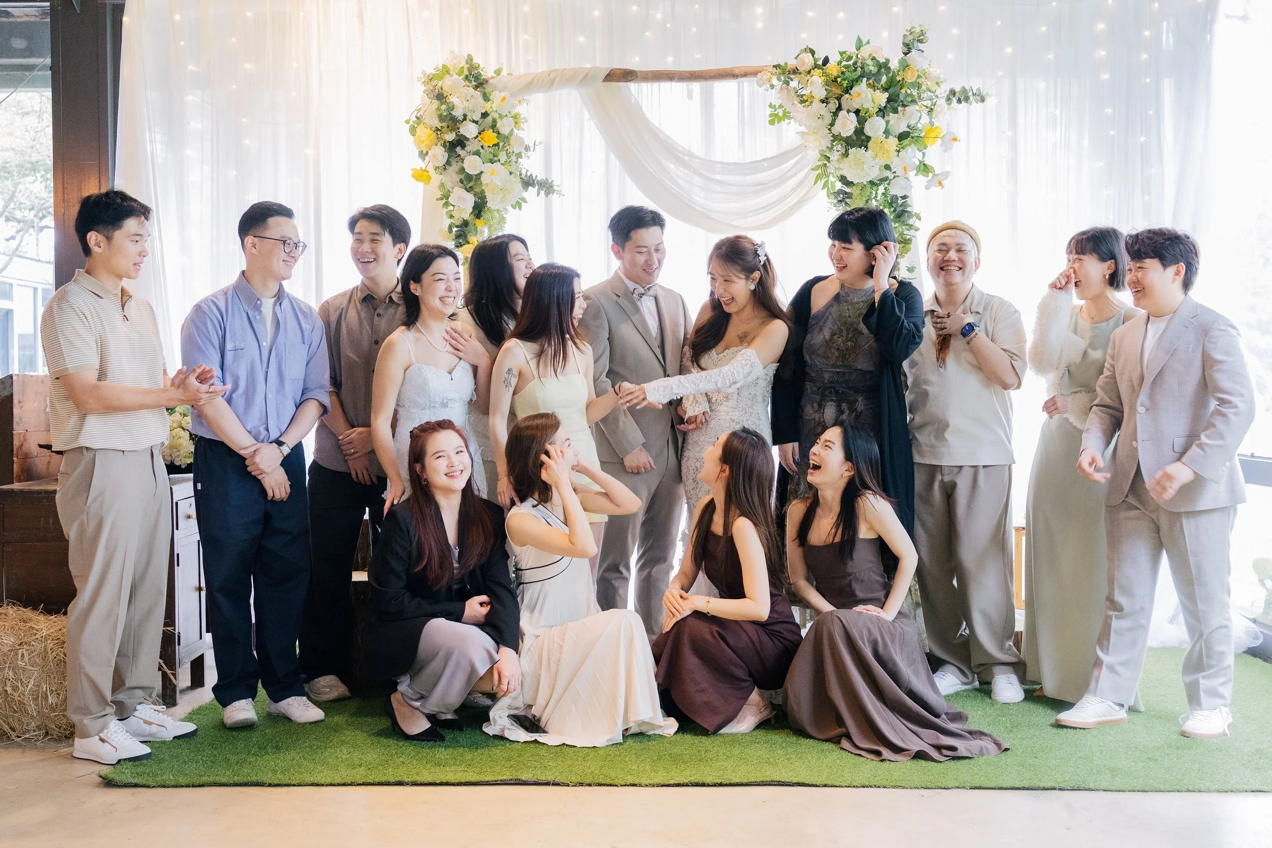 A group of people at a wedding celebration, standing and sitting in front of a decorated floral arch with white and yellow flowers, white drapery, and string lights in the background, smiling and laughing.