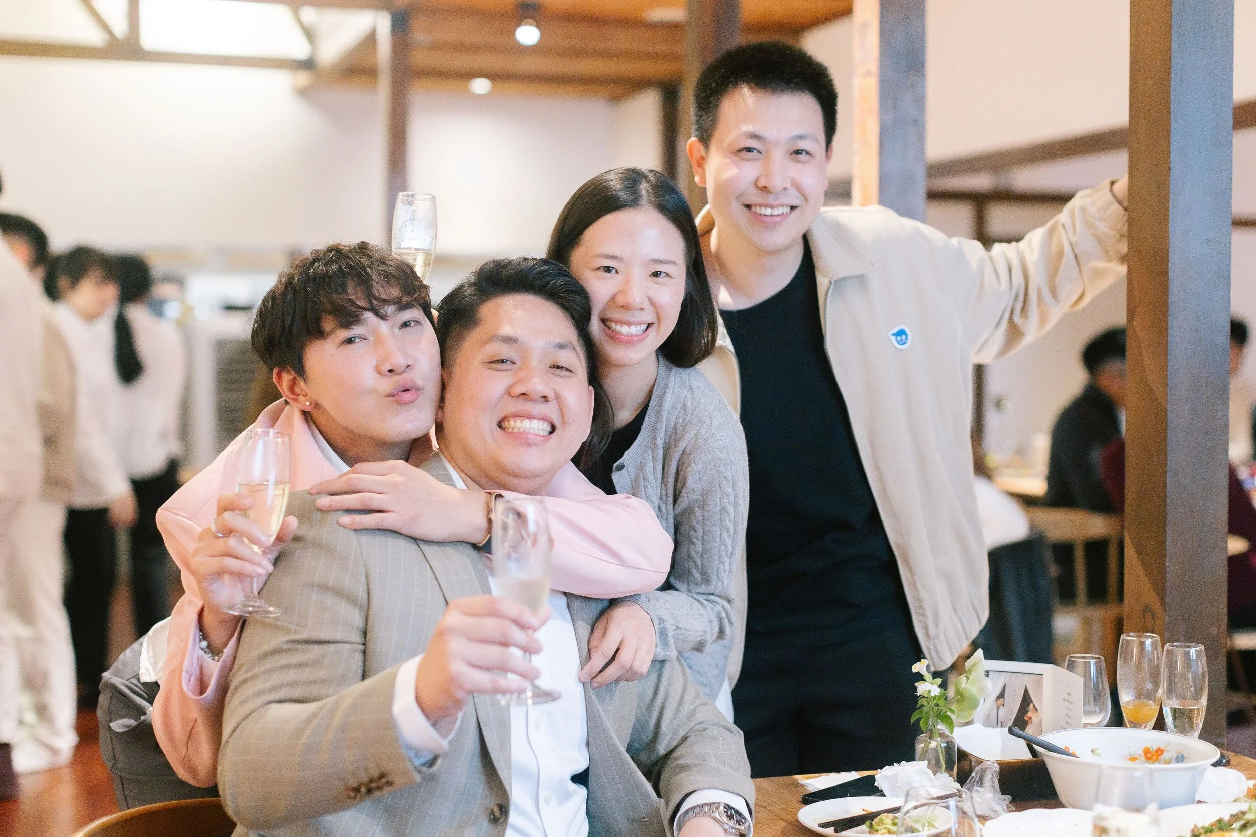 A group of five friends at a party, smiling, with some holding champagne glasses, sitting at a table with food and drinks, in a warmly lit indoor setting.