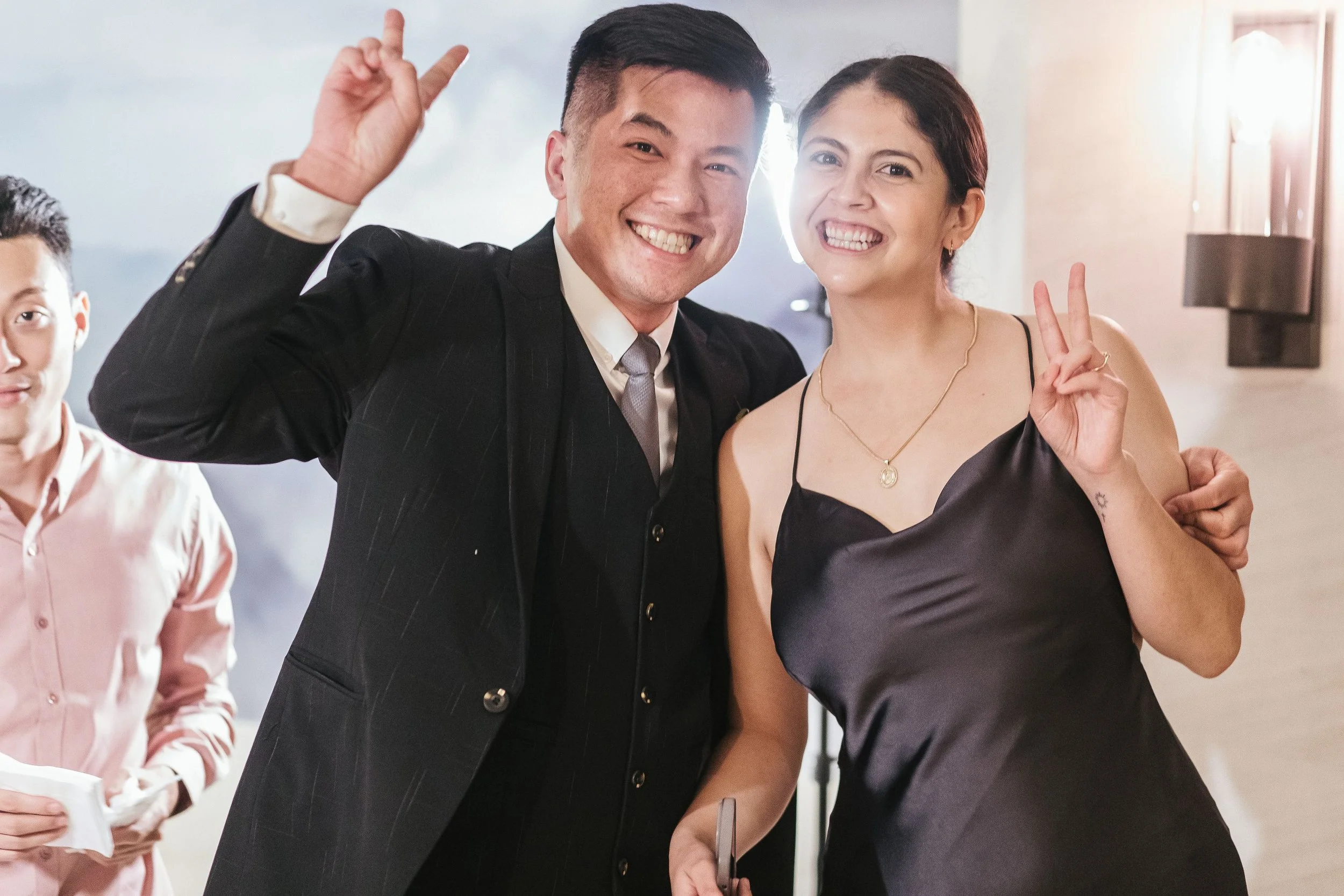 Group of people at a celebration or gathering, smiling and making peace signs, with sunlight in the background.