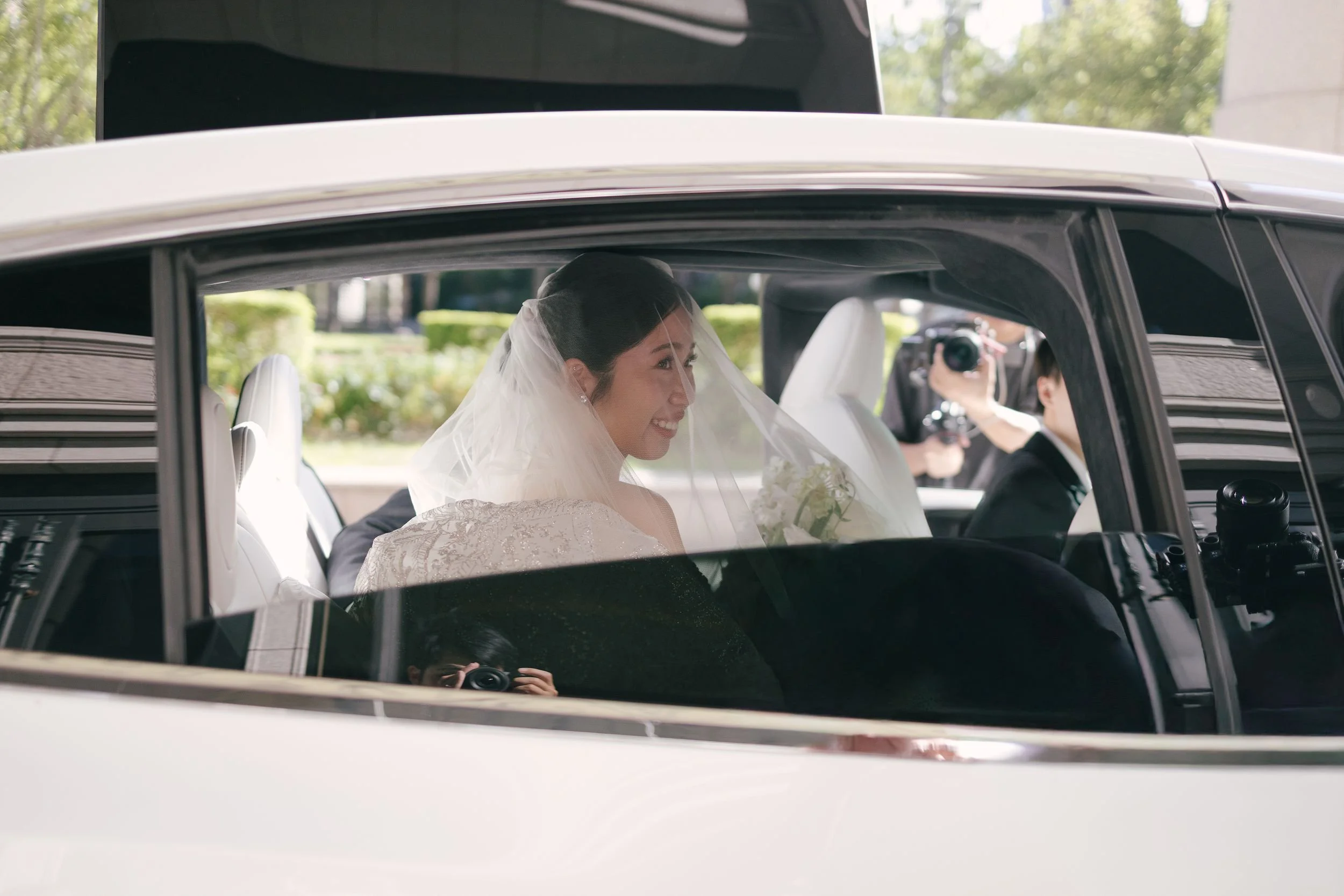 A bride with a veil smiling inside a luxury car, with someone taking a photo reflected in the car's window.