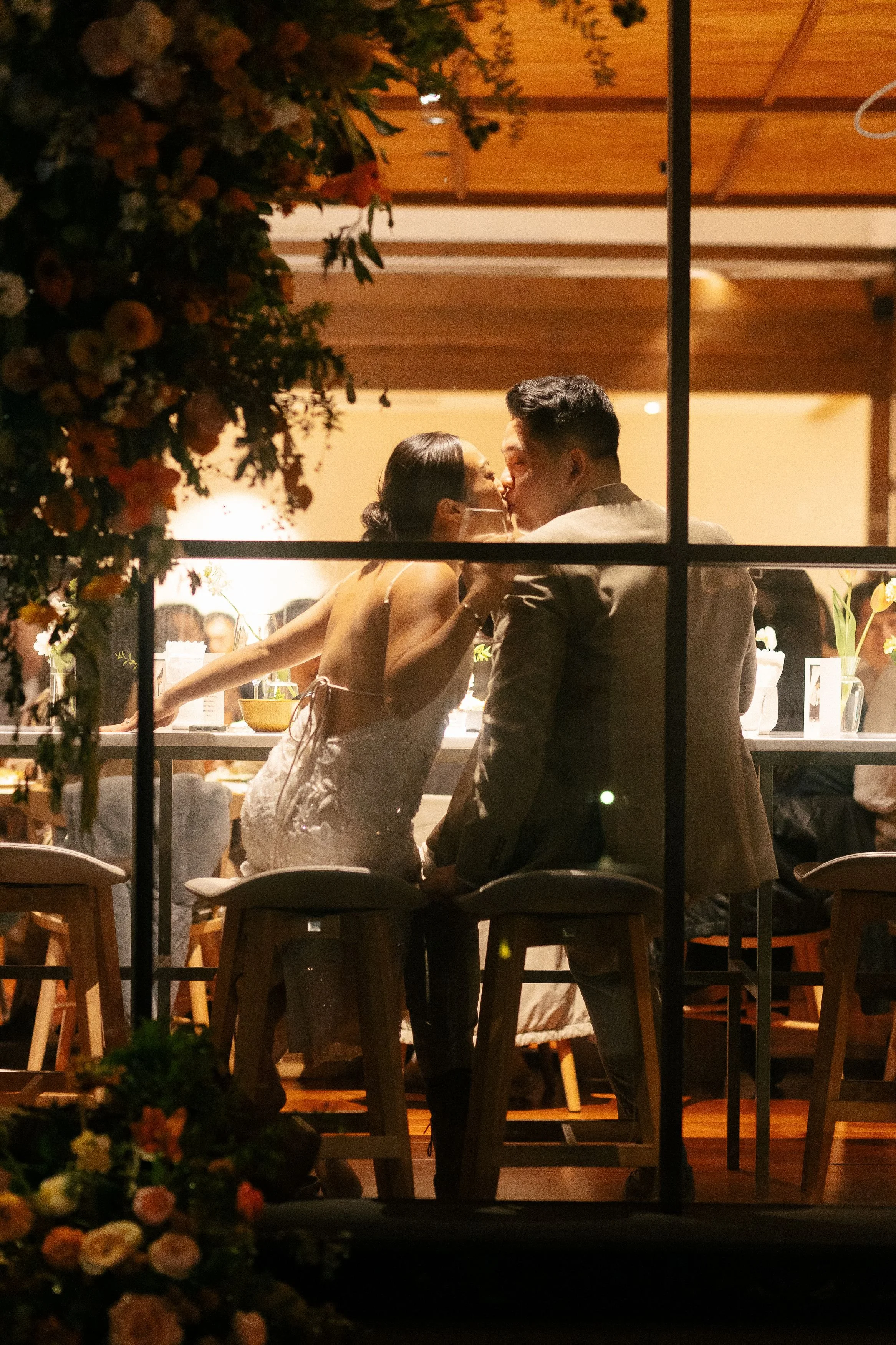 A couple is kissing inside a warmly lit room through a window, with floral decorations in the foreground.