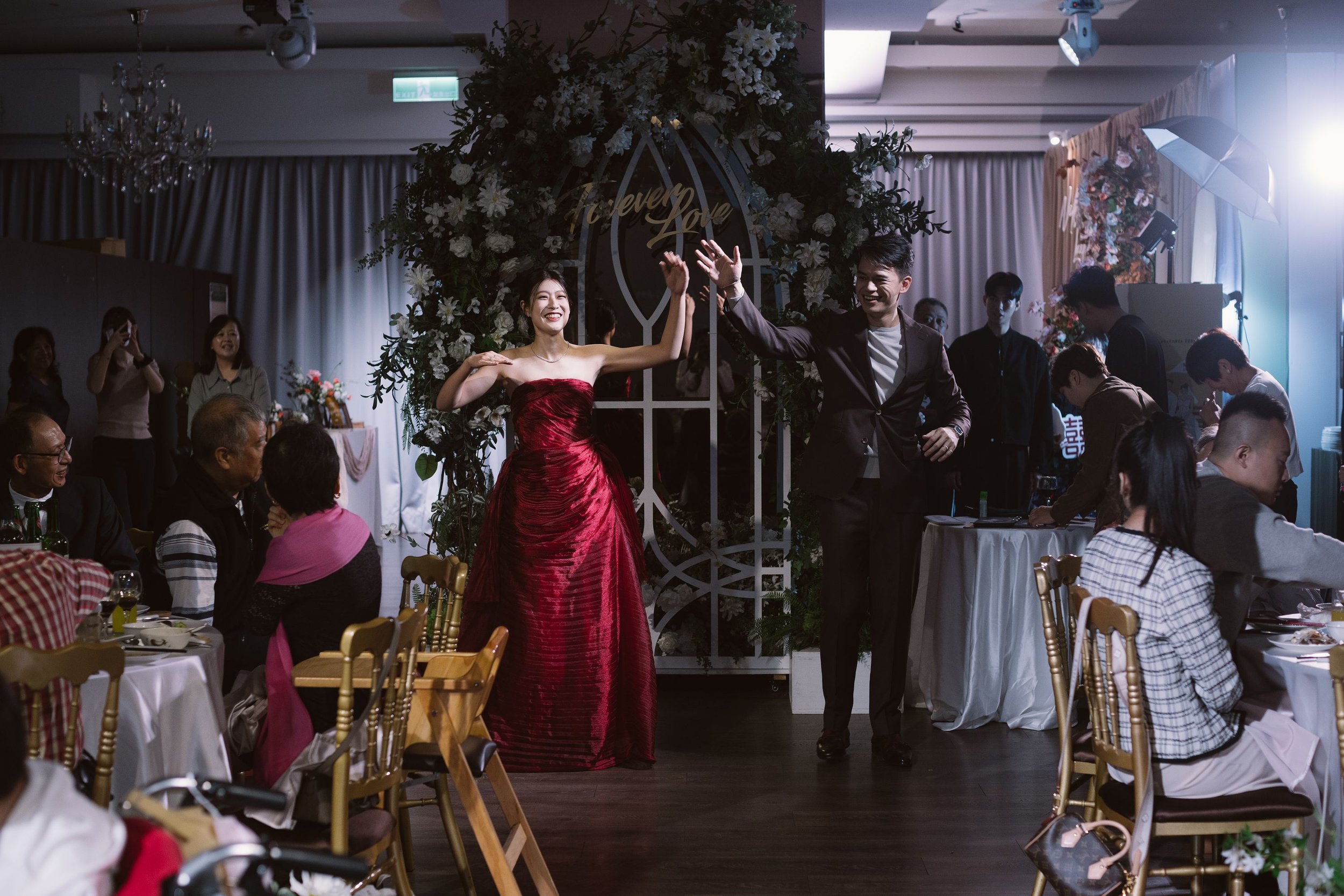 A woman in a red strapless gown and a man in a dark suit high-five during a celebration, with guests seated at decorated tables watching and taking photos. The setting appears to be a wedding or formal event, with floral arrangements and a decorative
