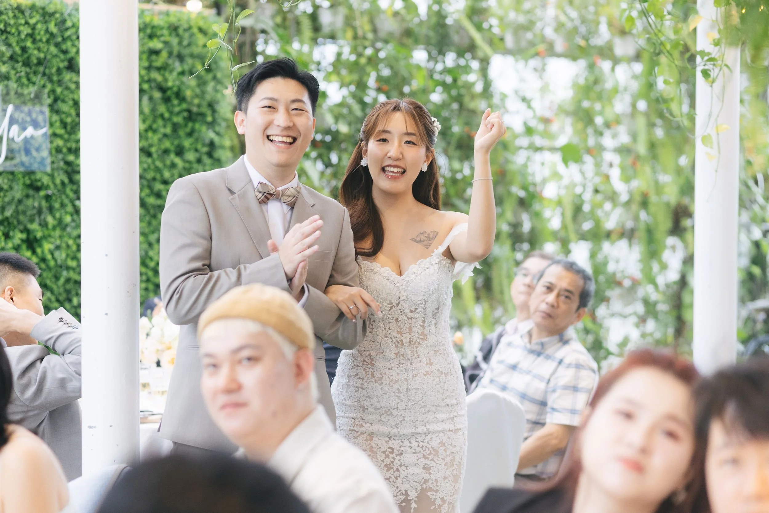 A smiling man in a light gray suit and a woman in a white lace dress, possibly at a wedding, standing outdoors with people seated around them, surrounded by greenery and hanging plants.