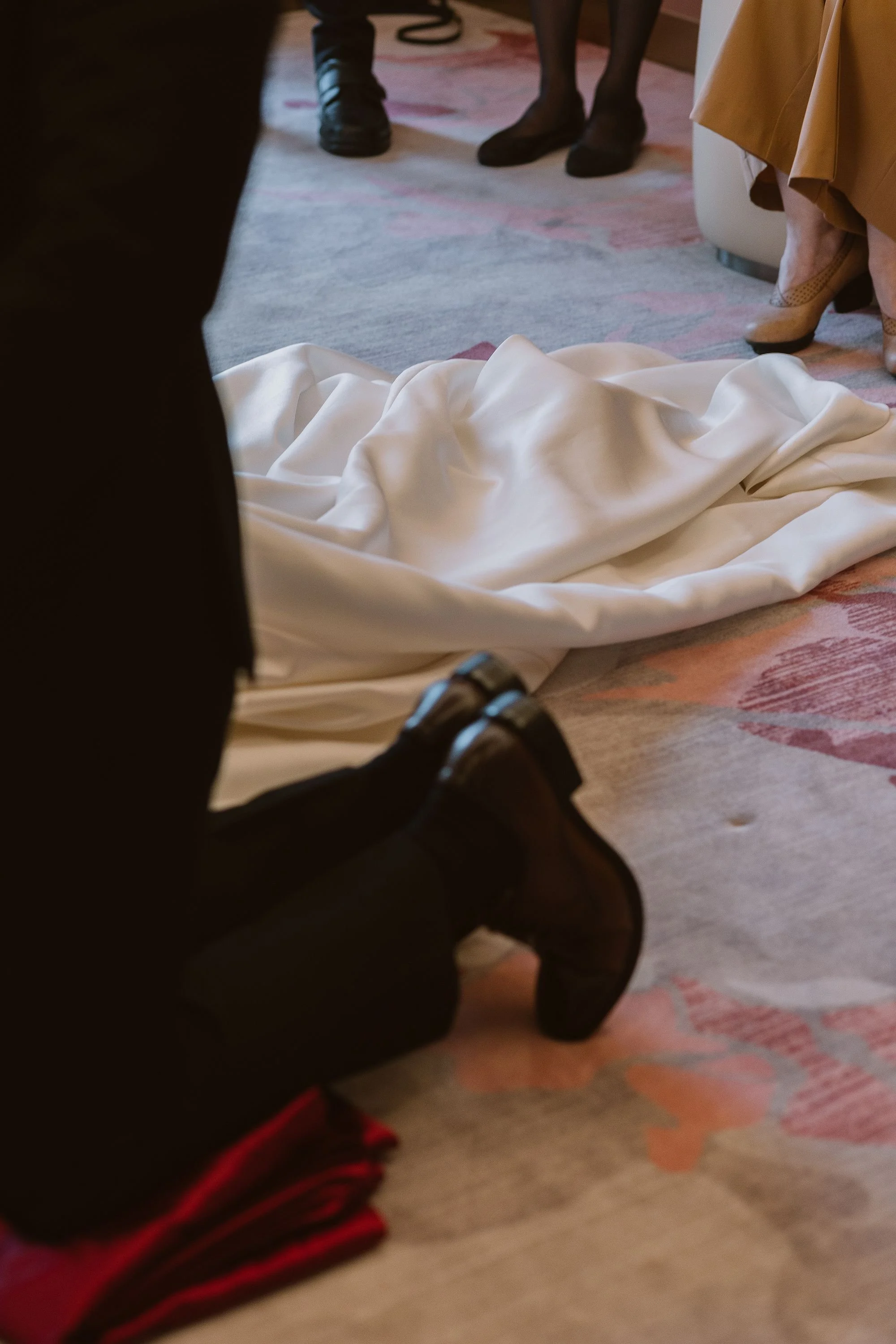 Close-up of people standing and sitting on a colorful carpet with a white fabric on the floor, possibly during a ceremony, with their feet visible.