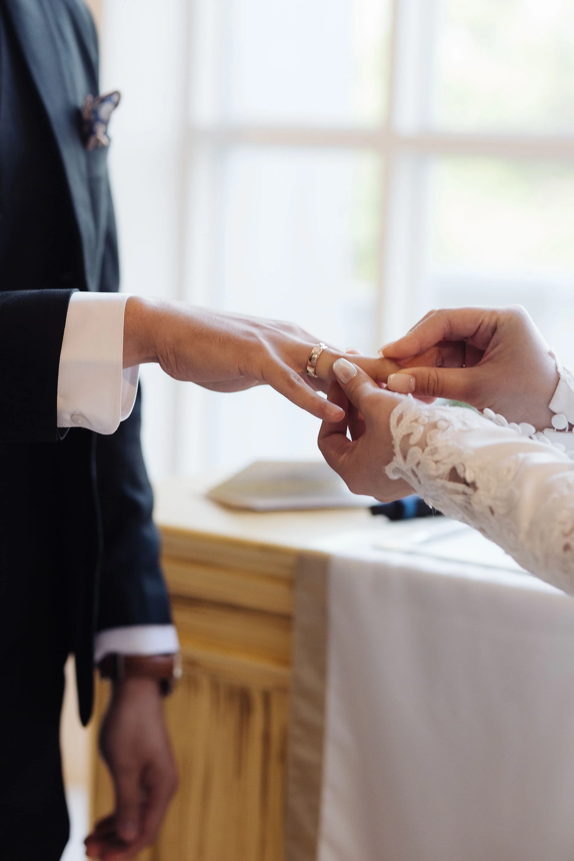 A person in a black suit is holding another person’s hand, who is wearing a white lace long sleeve dress, possibly during a wedding or ceremony. The person's torso and hand are visible, with a background of windows and wooden furniture.
