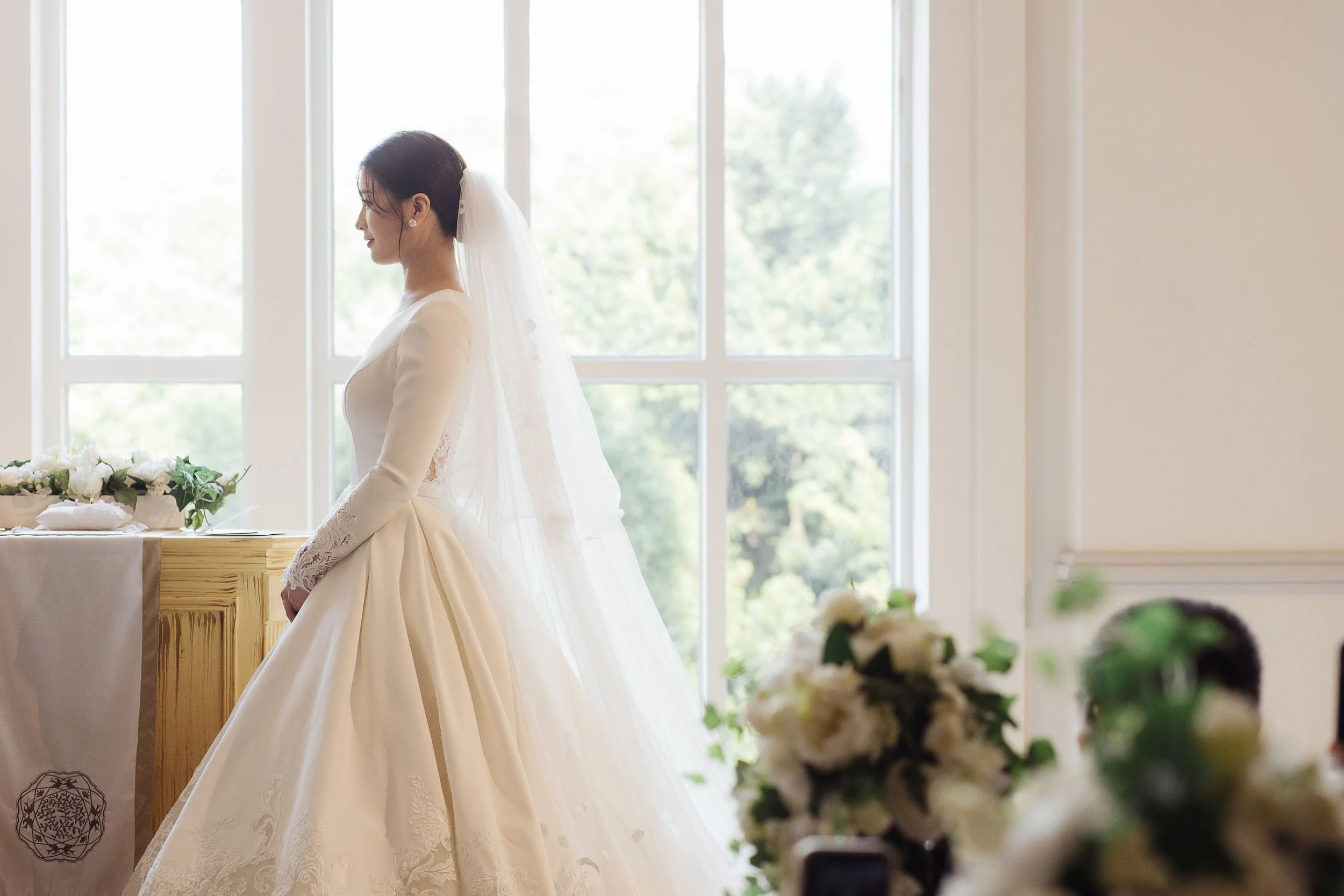 A bride in a white wedding dress with veil standing indoors in front of large windows, with a table decorated with flowers and greenery nearby.