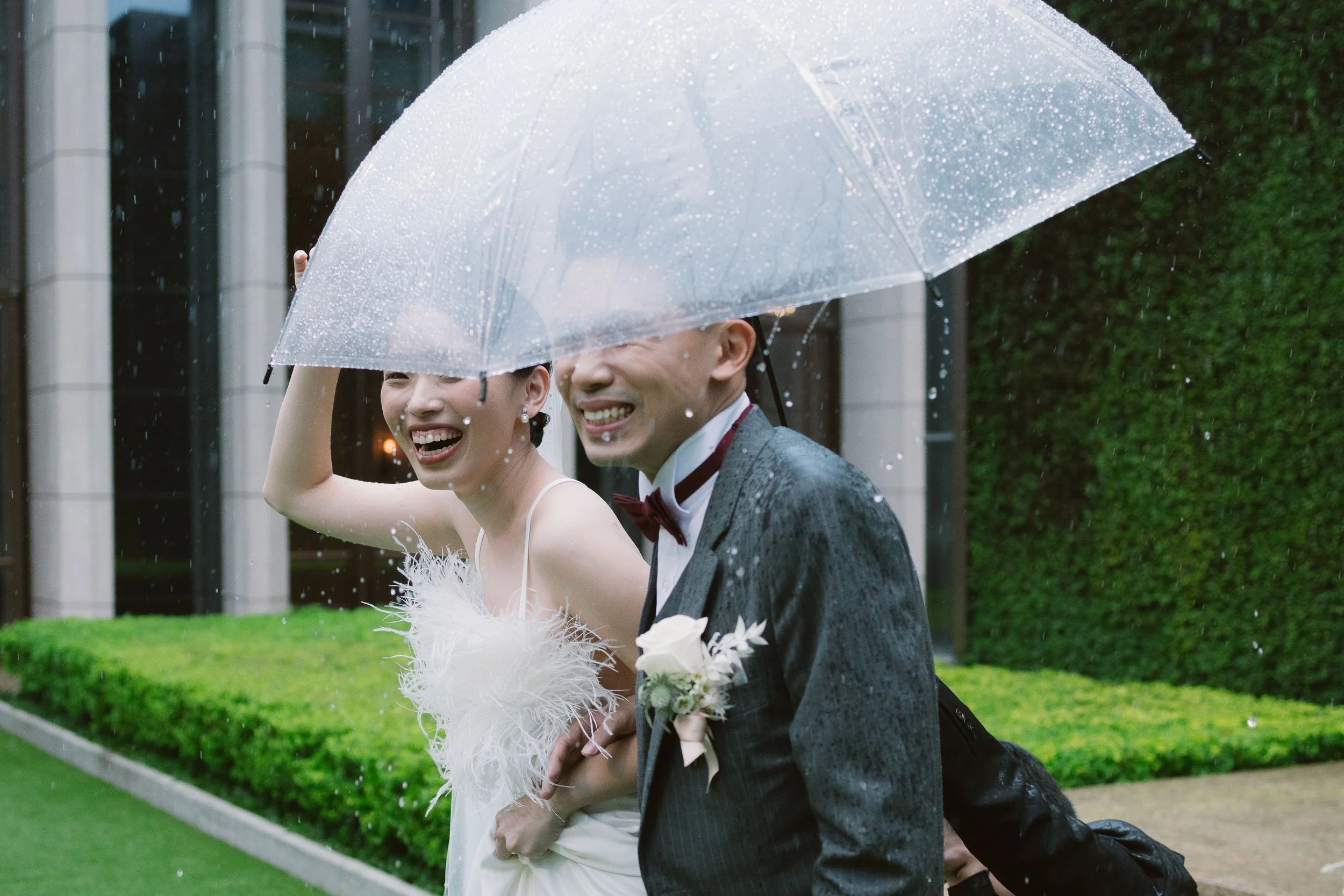 A happy bride and groom walking in the rain under a transparent umbrella, smiling and enjoying their wedding day.