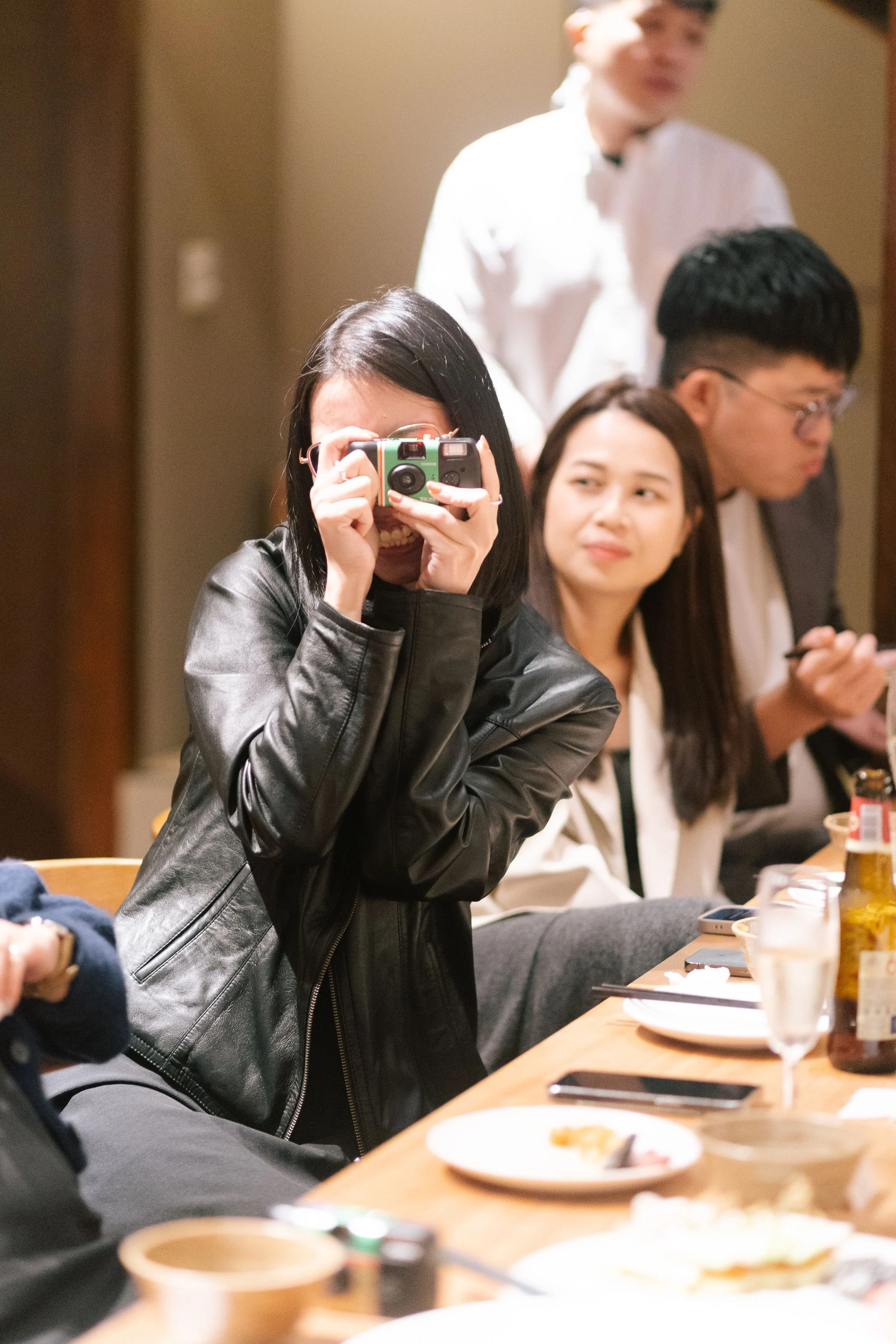 A woman takes a photo with a camera, smiling, at a social gathering. Other people sit around a table with food and drinks, engaged in conversation.
