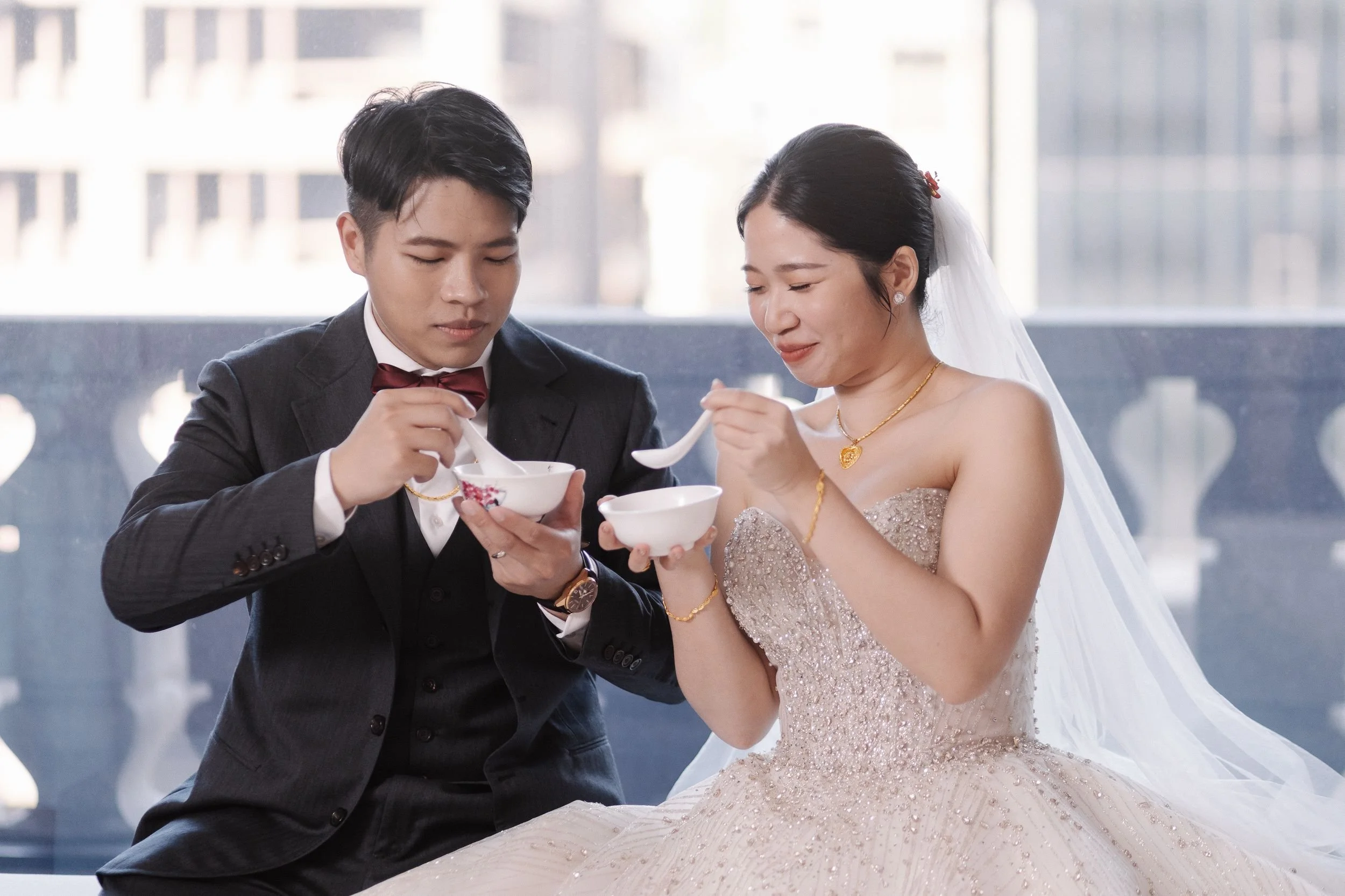 A bride and groom dressed in wedding attire sharing a meal with spoons during their wedding celebration, sitting close together indoors with a cityscape view in the background.