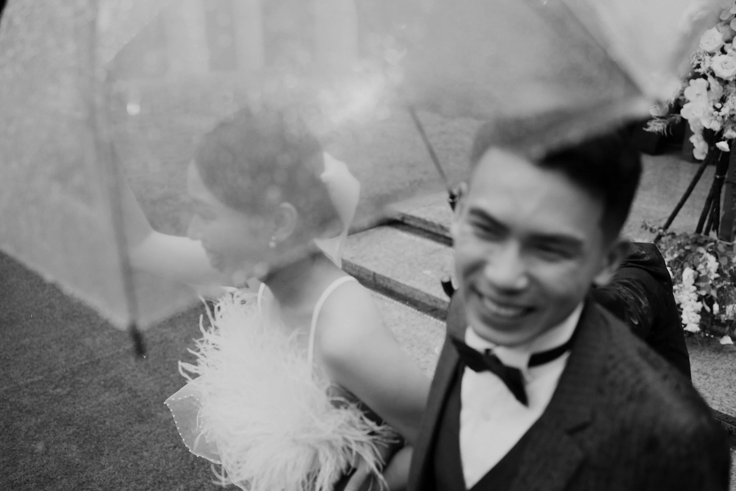 A young boy dressed in formal attire, including a tuxedo and bow tie, bowing with a big smile. He is sitting on the ground near a decorated, furry, stuffed animal on a table with floral arrangements. The setting appears to be a celebratory event or c