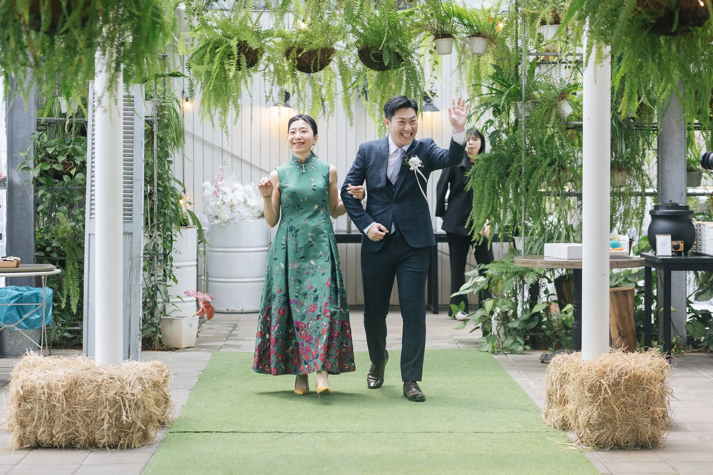 Smiling couple walking down a green carpet in a greenhouse decorated with hanging plants, surrounded by hay bales.