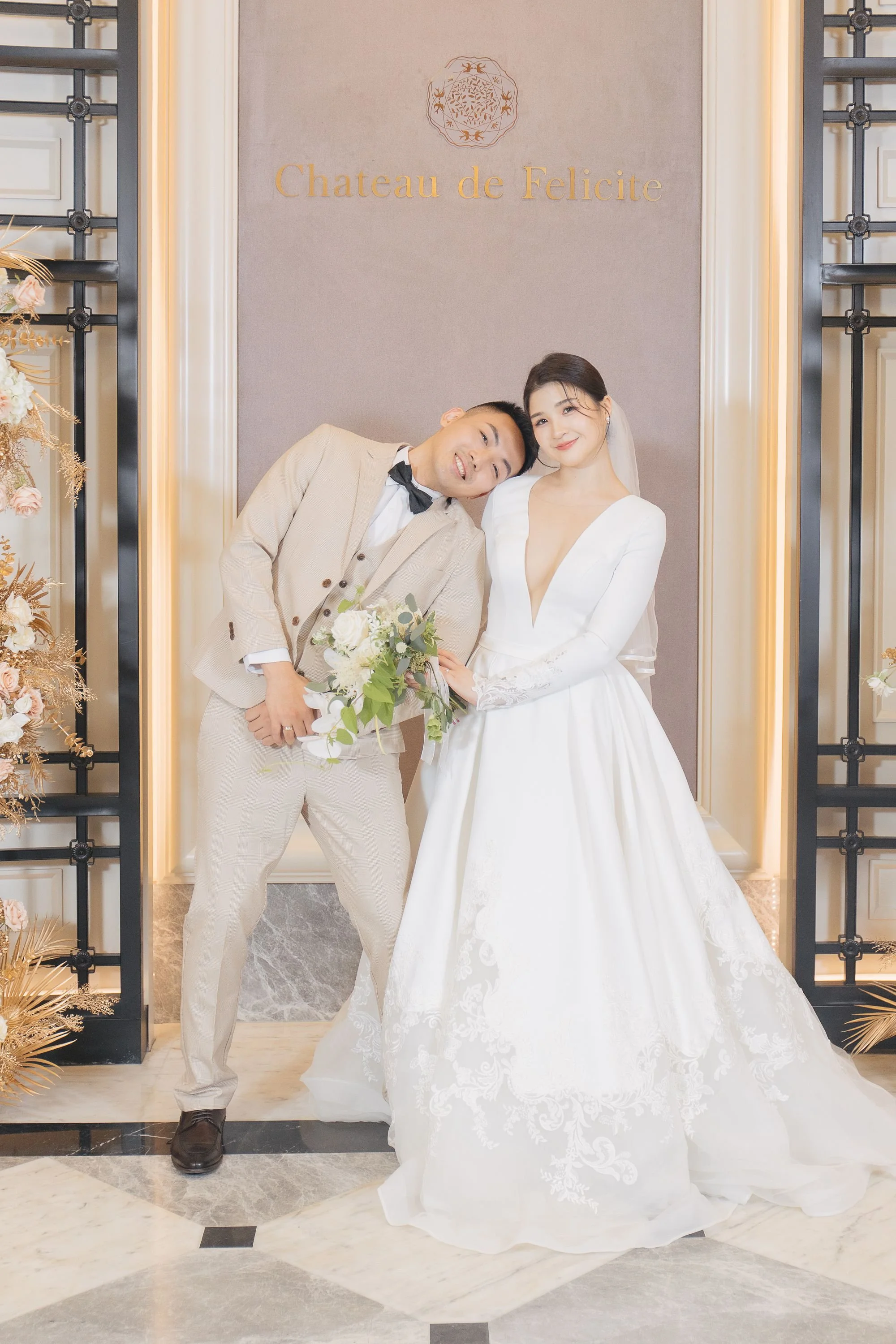 A bride and groom posing together at their wedding, standing in front of a wall with the sign 'Chateau de Felicite.' The bride wears a white gown with a deep V neckline and lace details, and the groom wears a beige checked suit with a black bow tie. 