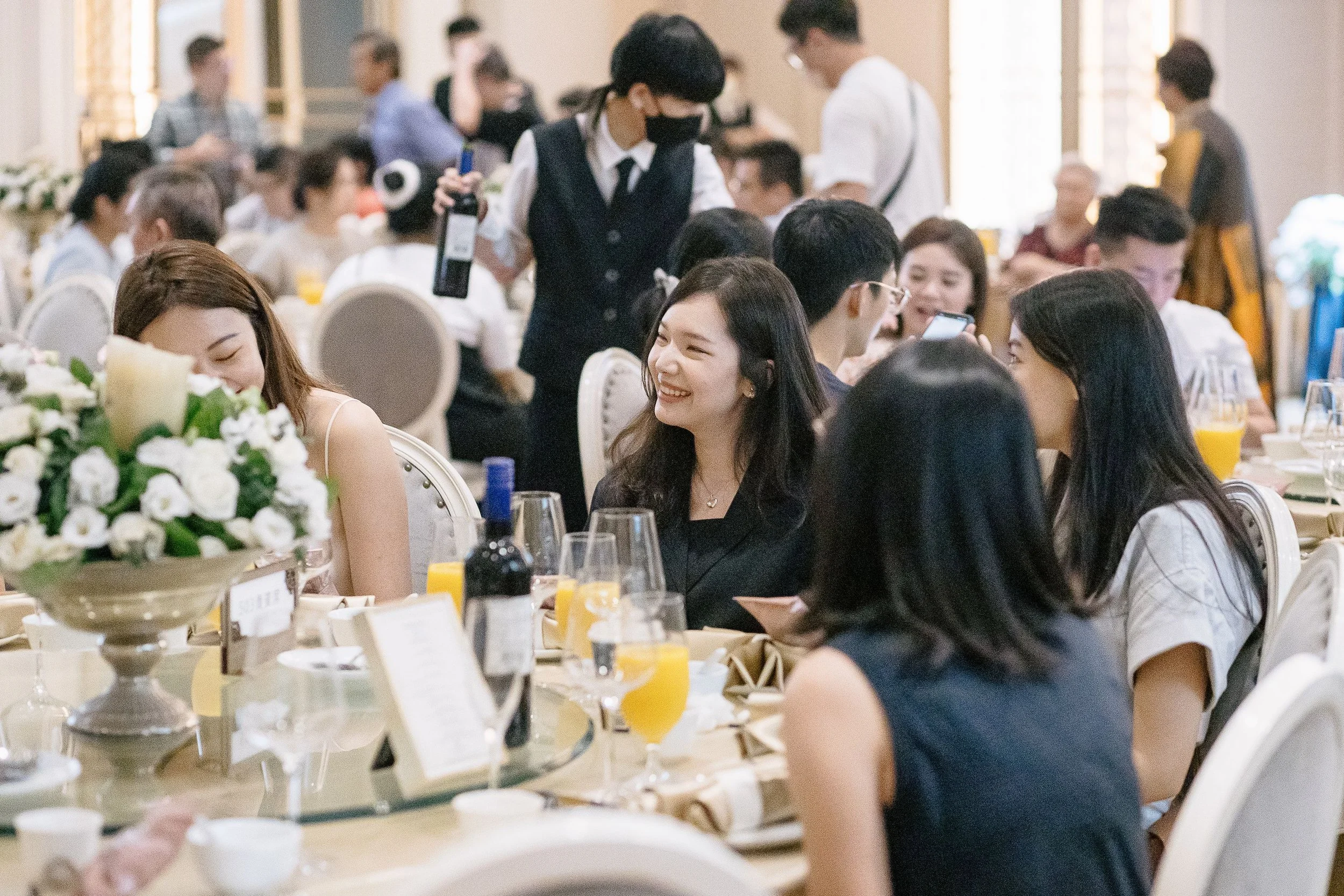 A group of people gathered at a banquet, sitting at tables with floral centerpieces and food, with a waiter serving in the background.