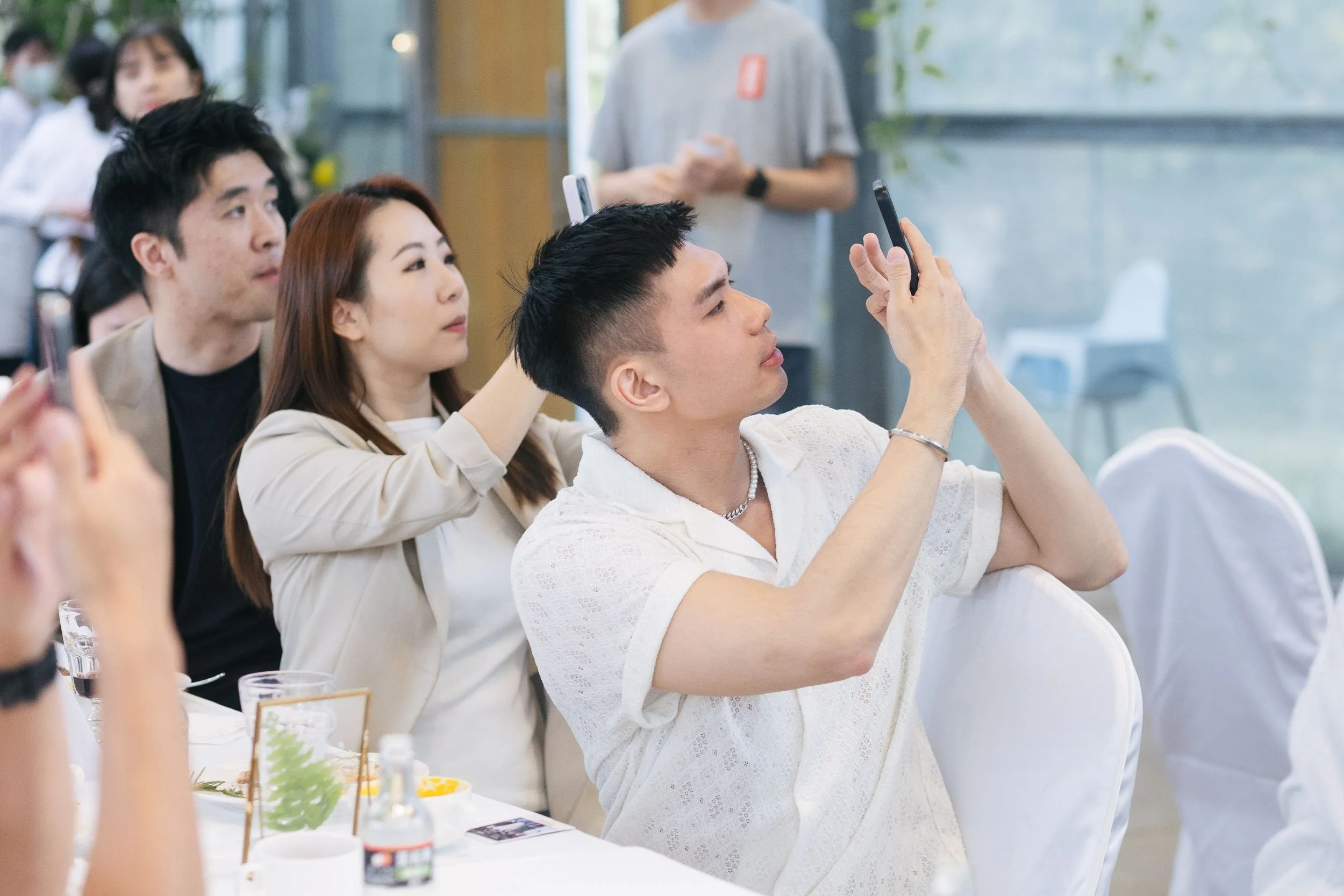A group of people sitting at a table during an event, with one person taking a photo or video with a smartphone. The setting appears to be indoors, in a bright, modern space.