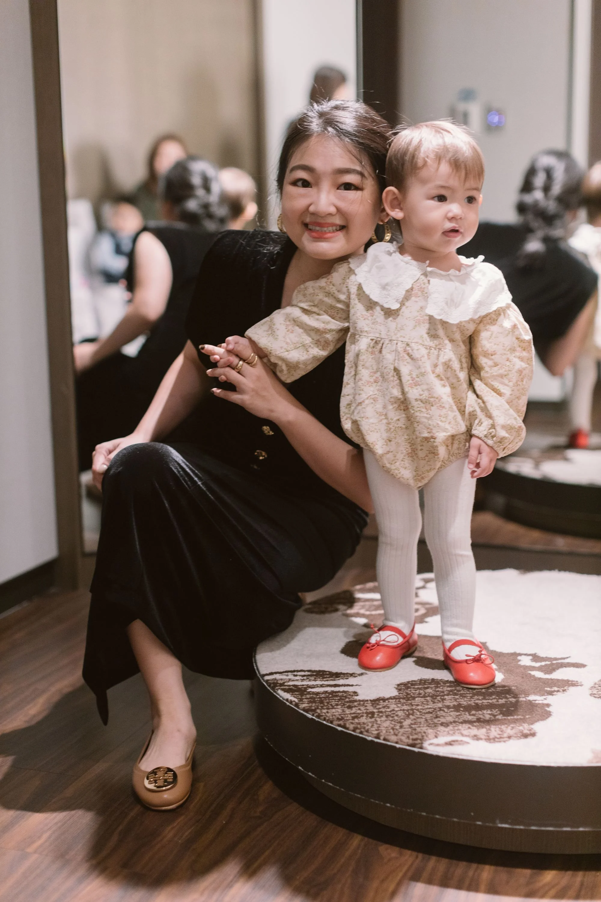 A woman with dark hair smiling while holding a young girl with light brown hair, dressed in a floral dress, white tights, and red shoes, standing on a round, patterned rug in a room filled with other people.