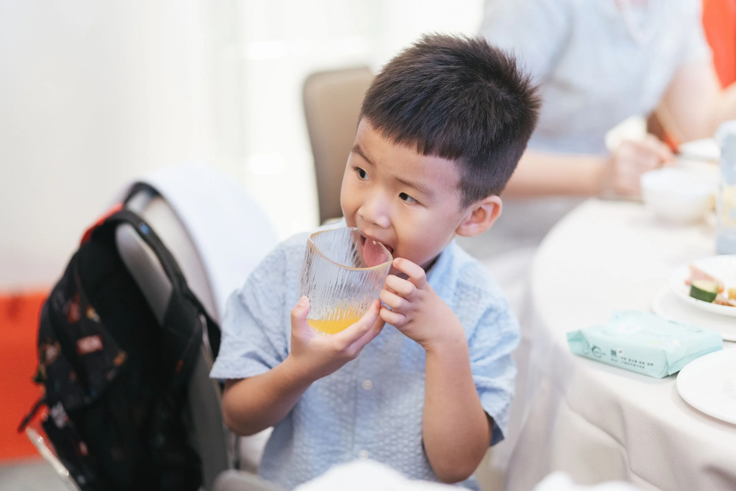 A young boy with short black hair, wearing a light gray shirt, drinks from a glass of yellow-orange beverage, sitting at a table with a white tablecloth surrounded by plates, bowls, and food items.