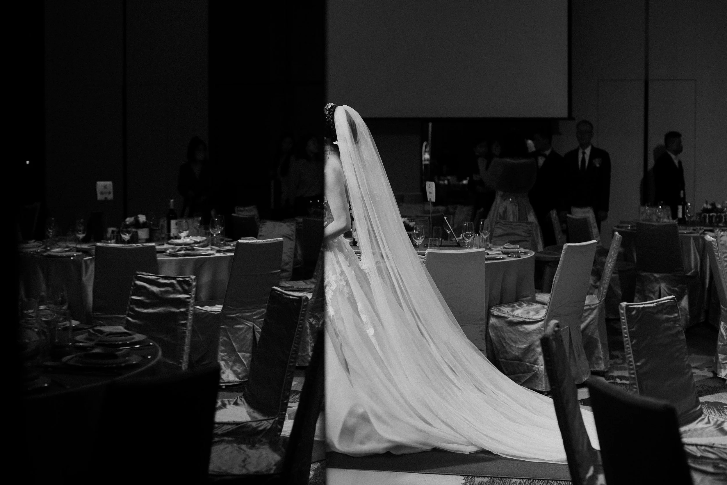 A bride in a wedding gown and veil standing at a wedding reception, surrounded by guests and decorated tables.