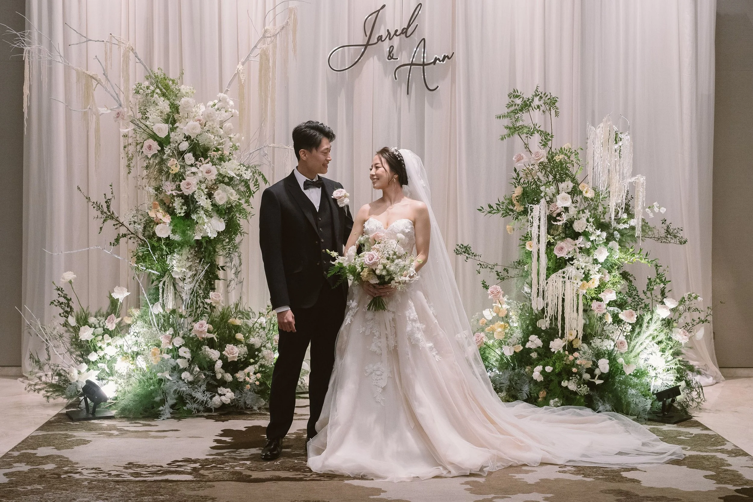 A bride and groom standing together at their wedding, surrounded by floral arrangements, with a backdrop featuring the names Jared and Ann.