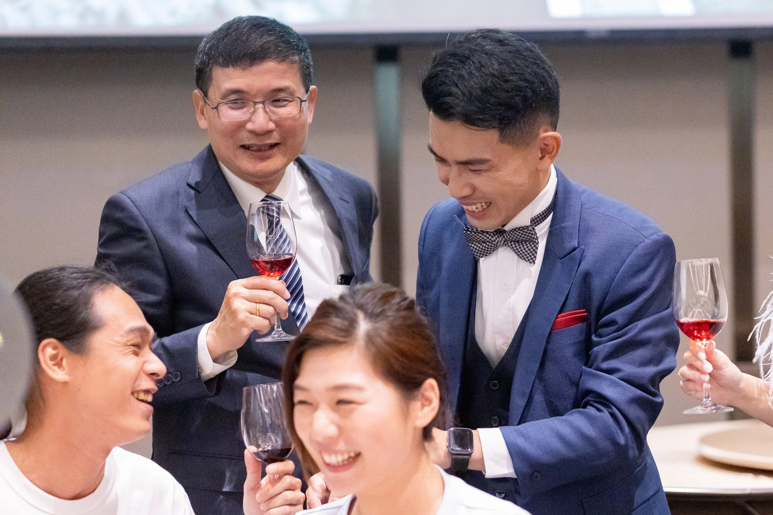 Two men in formal attire, one with glasses holding a glass of red wine, smiling and engaging with others at a social gathering.