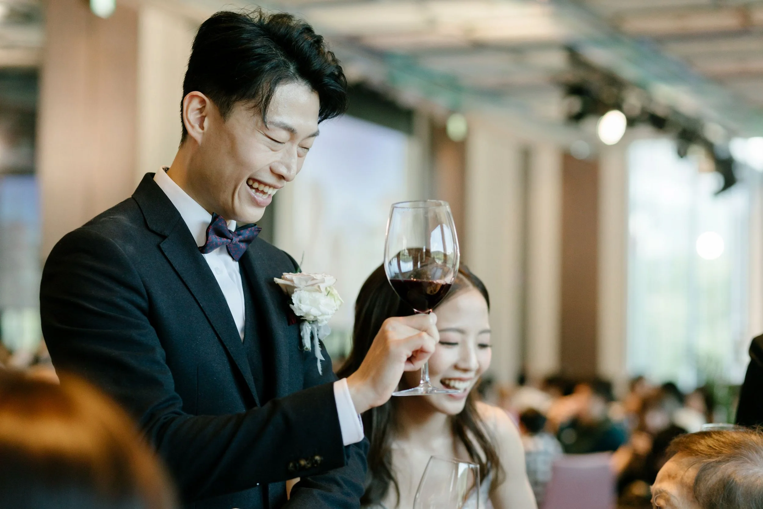 A man in a tuxedo and bow tie holding a glass of red wine, smiling at a social gathering.