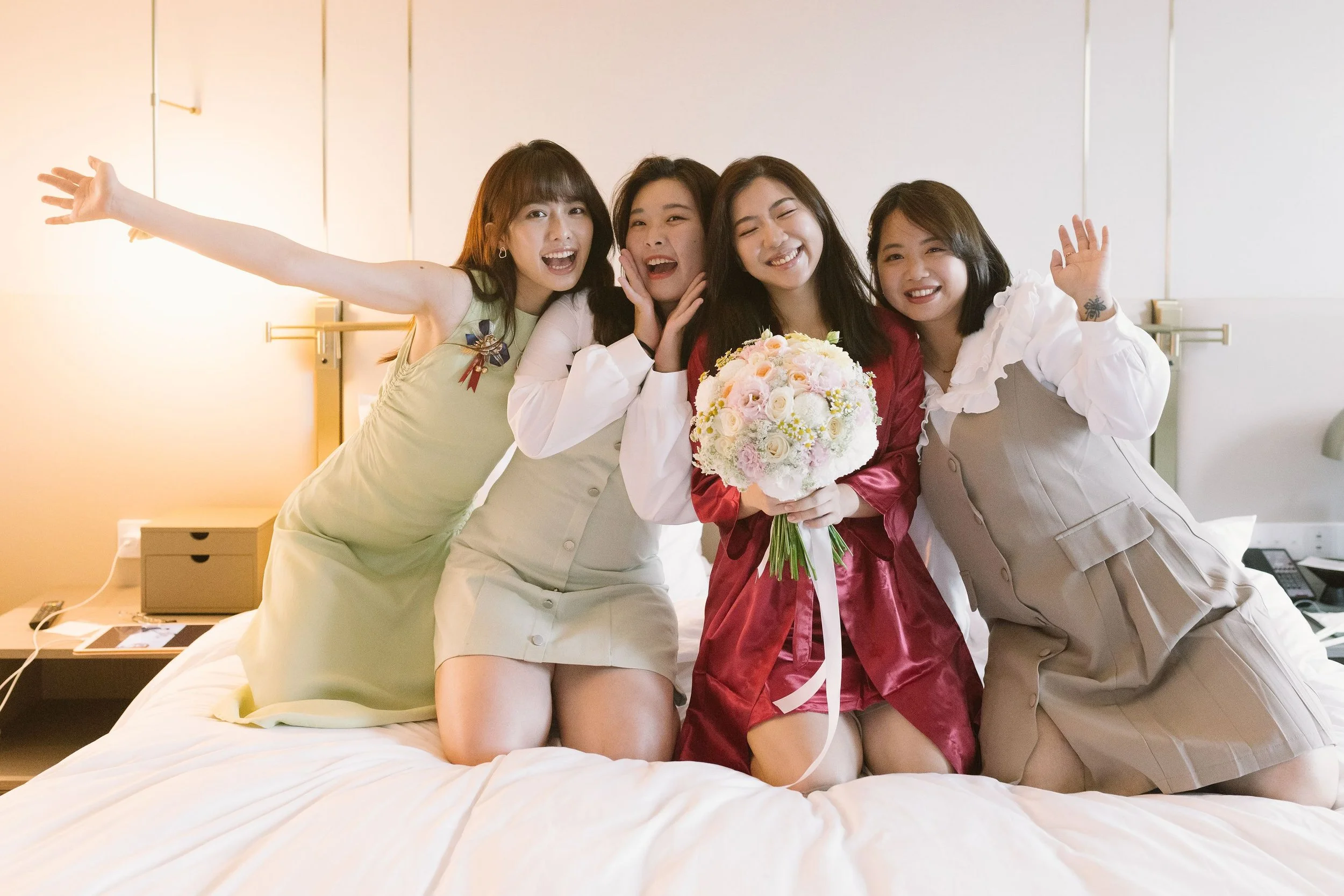 Four women sitting on a bed, celebrating with a bouquet of flowers, smiling, and waving. One woman in the center is wearing a red satin robe, while the others wear pastel dresses. The background is a neutral-colored bedroom with a nightstand and some
