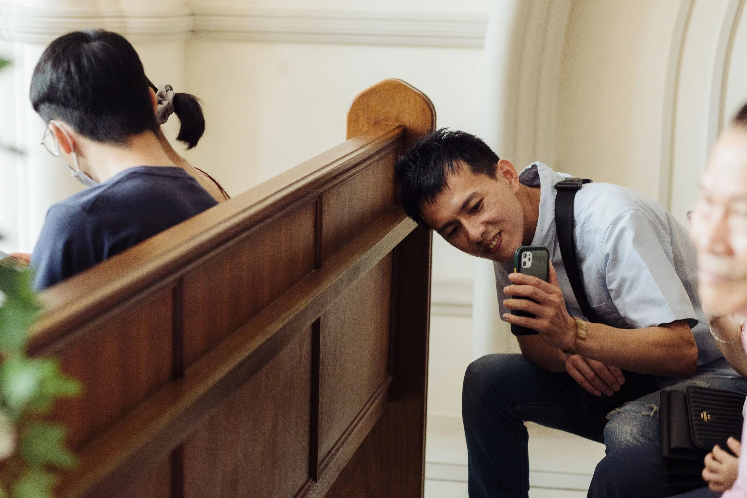 A group of people, including a young man holding a smartphone, sitting and leaning on a wooden divider in an indoor setting.