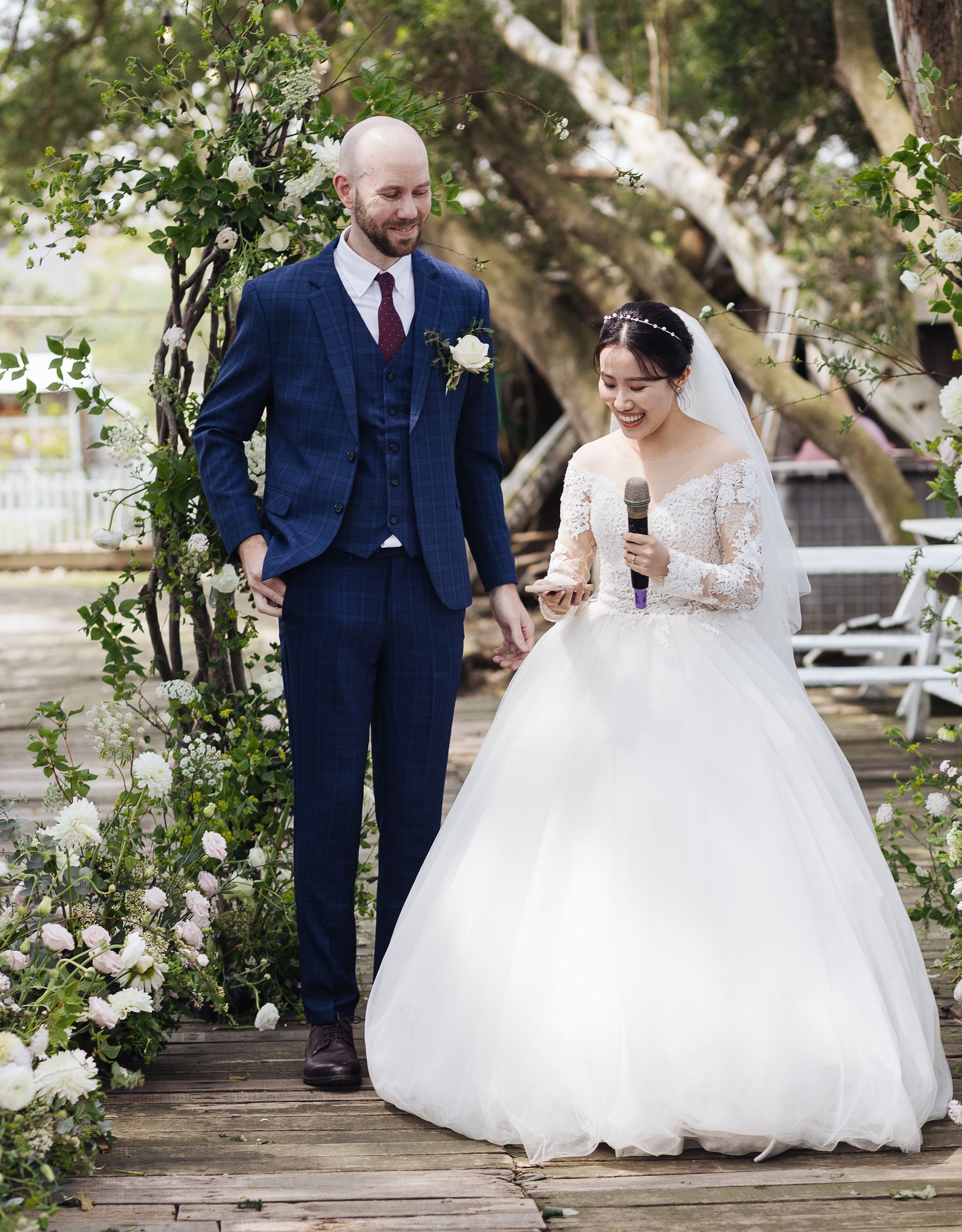 A bride and groom holding hands during their wedding ceremony outdoors, surrounded by floral arrangements and greenery, with the bride reading from a phone and holding a microphone.