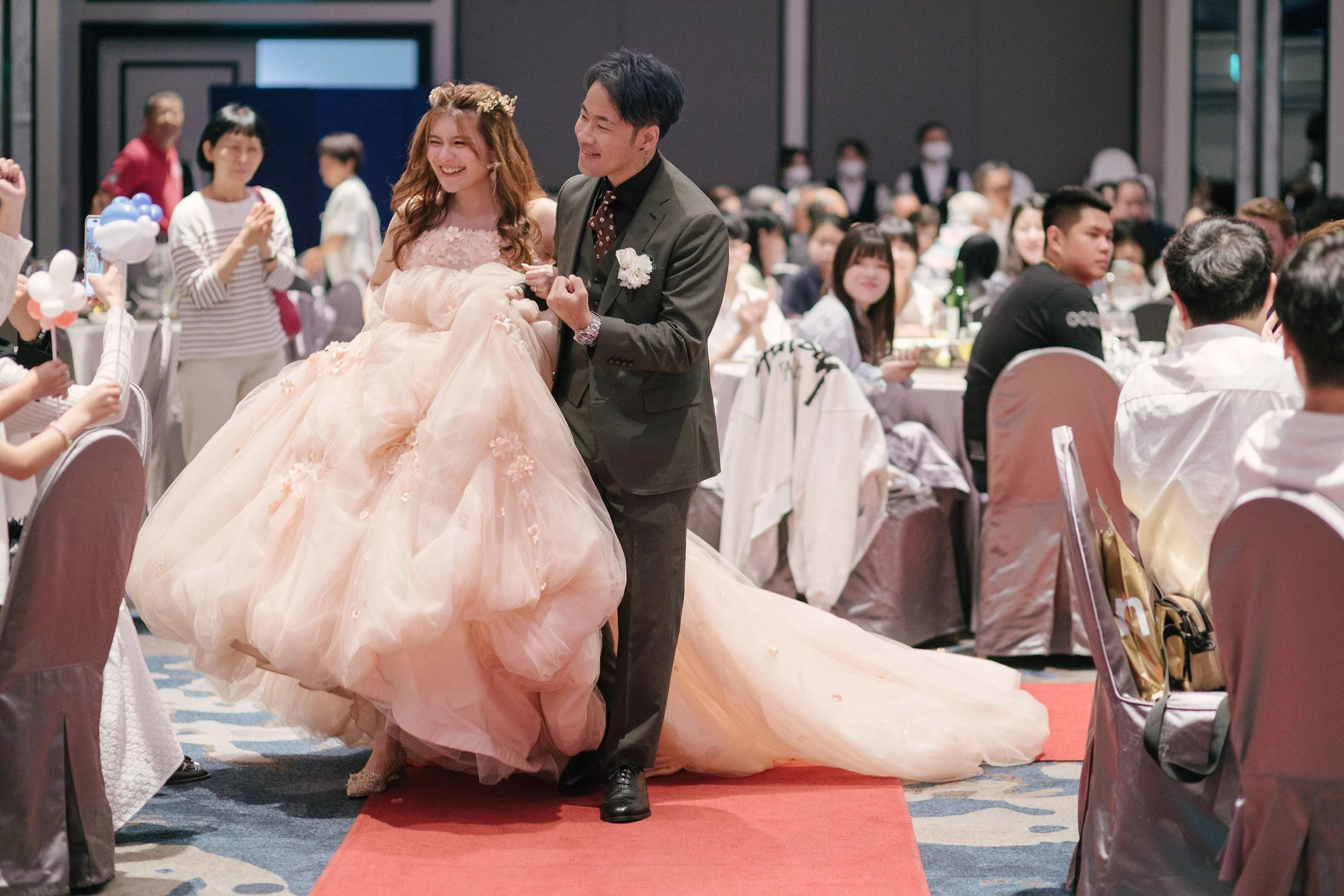 A bride in a voluminous peach-colored wedding gown with floral details and a groom in a dark suit, smiling and holding hands, at a wedding reception with seated guests and decorations in the background.